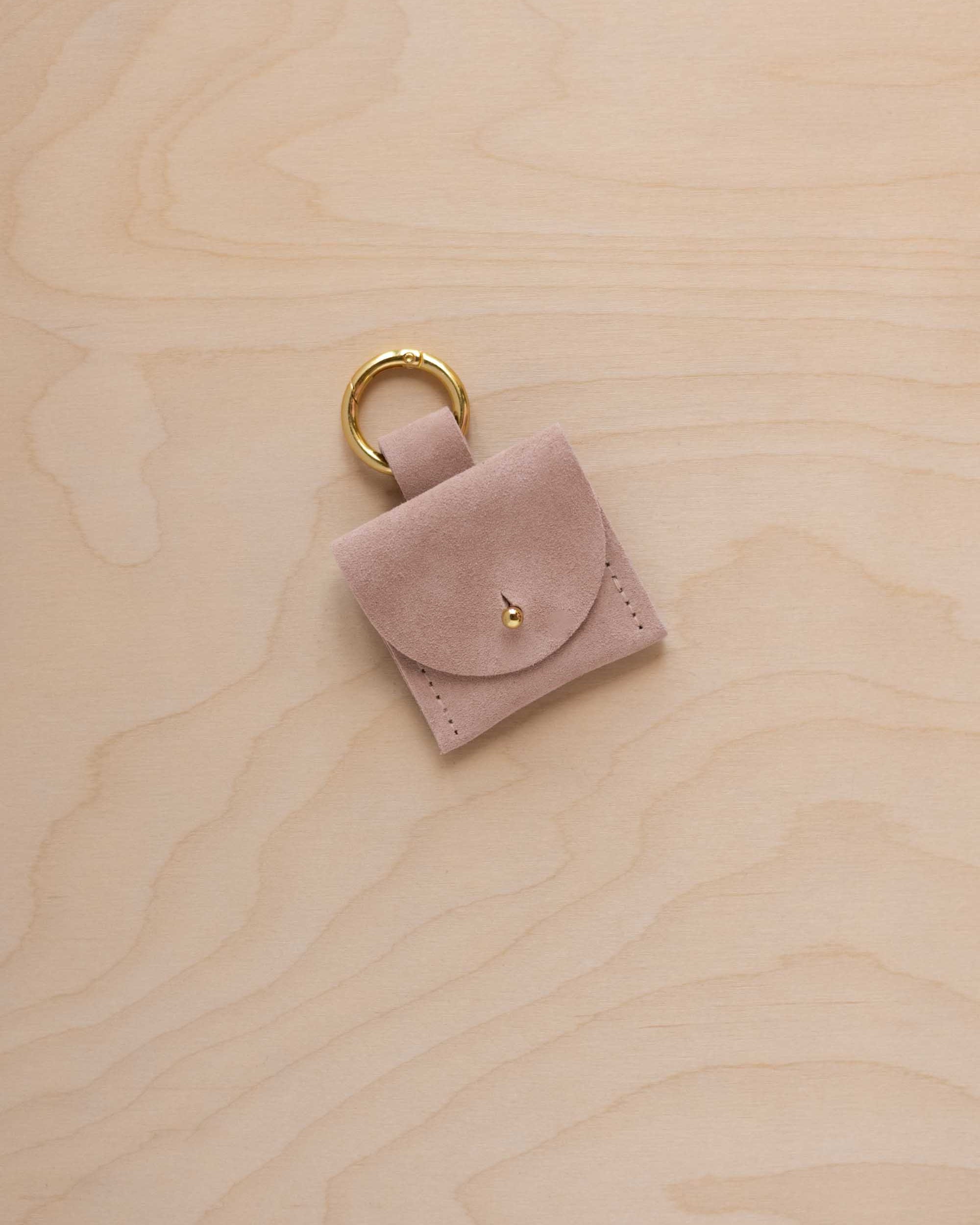 A small beige suede pouch bag charm with a gold coloured clip ring fastening, displayed on a light wood grain background.