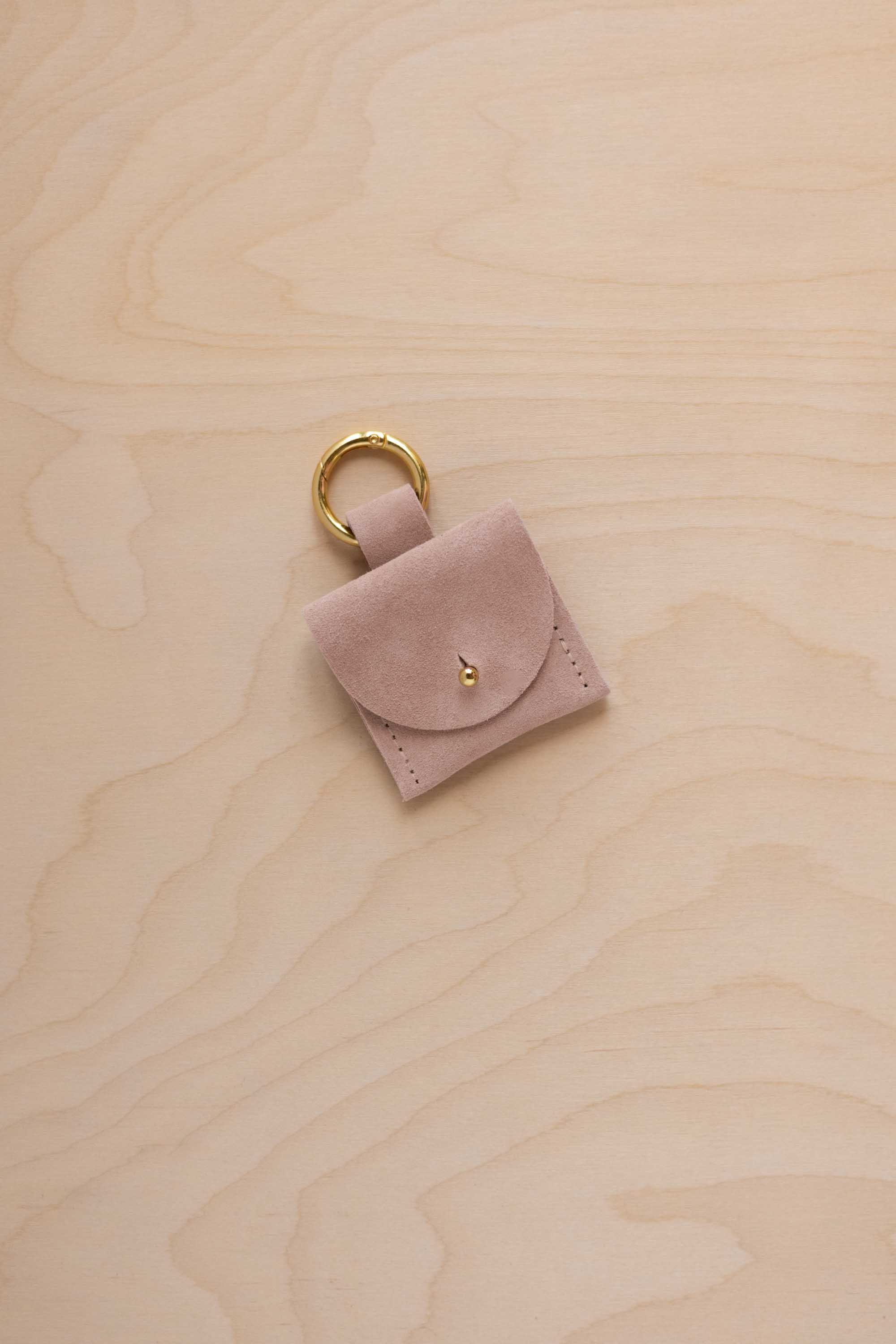 A small beige suede pouch bag charm with a gold coloured clip ring fastening, displayed on a light wood grain background.