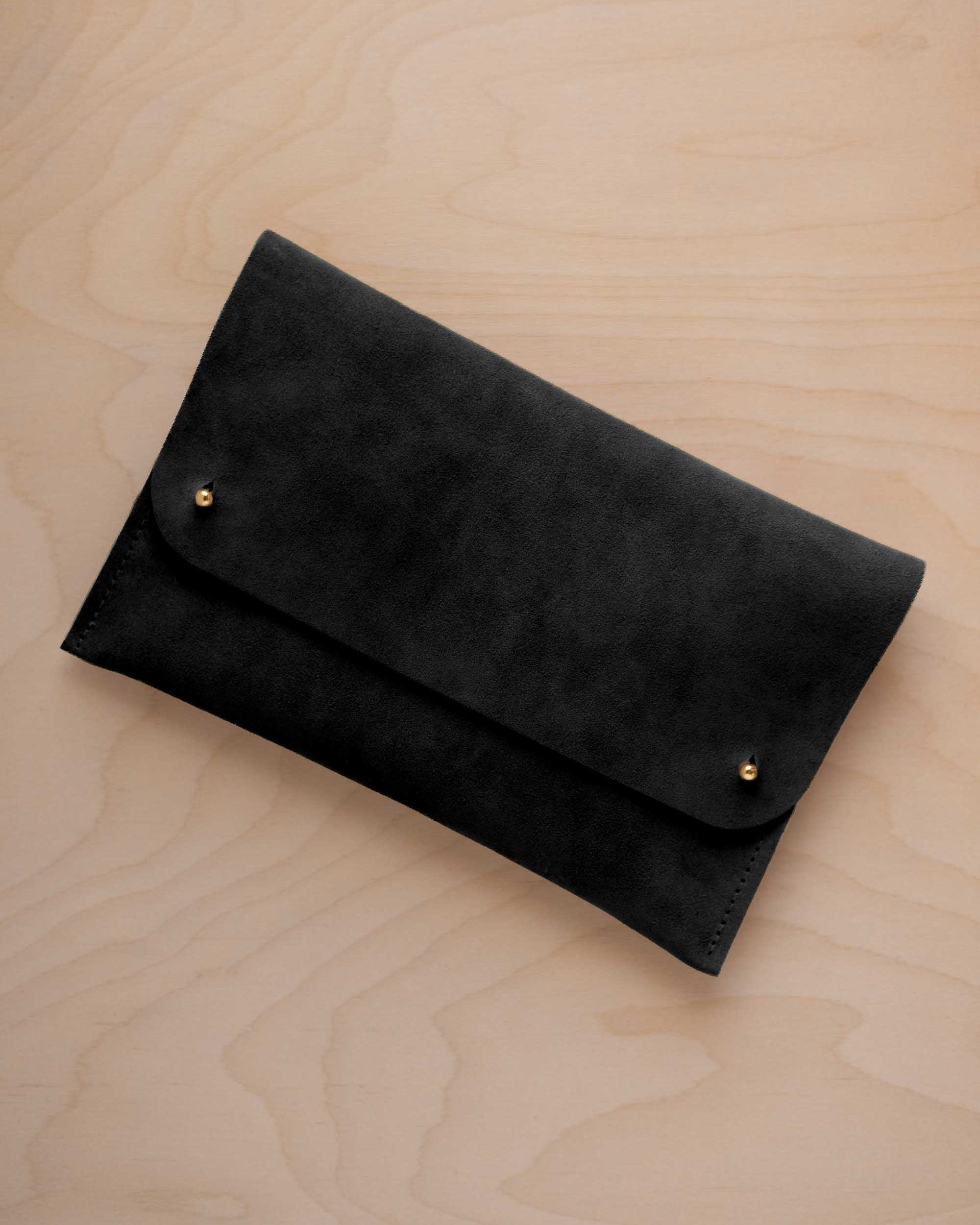 A black suede clutch with small gold coloured metal studs, displayed diagonally on a light wood grain surface.