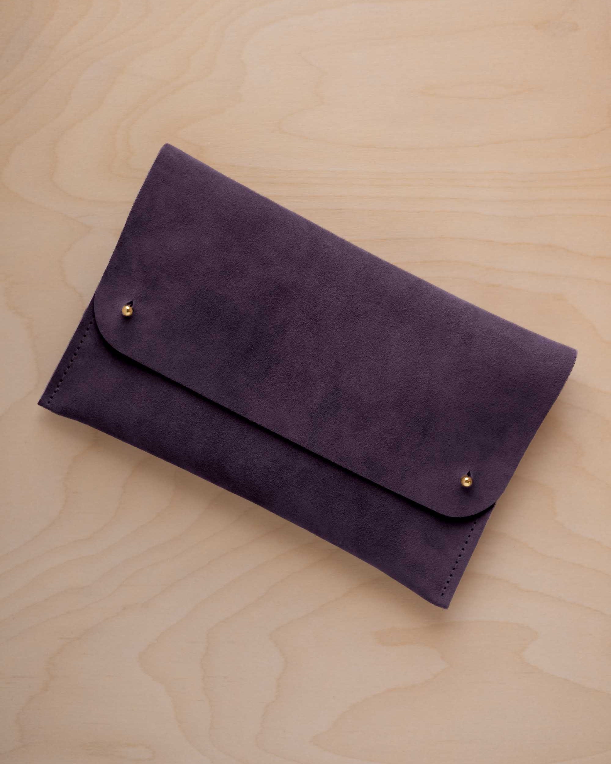 A blackcurrant dark purple suede clutch with small gold coloured metal studs, displayed diagonally on a light wood grain surface.