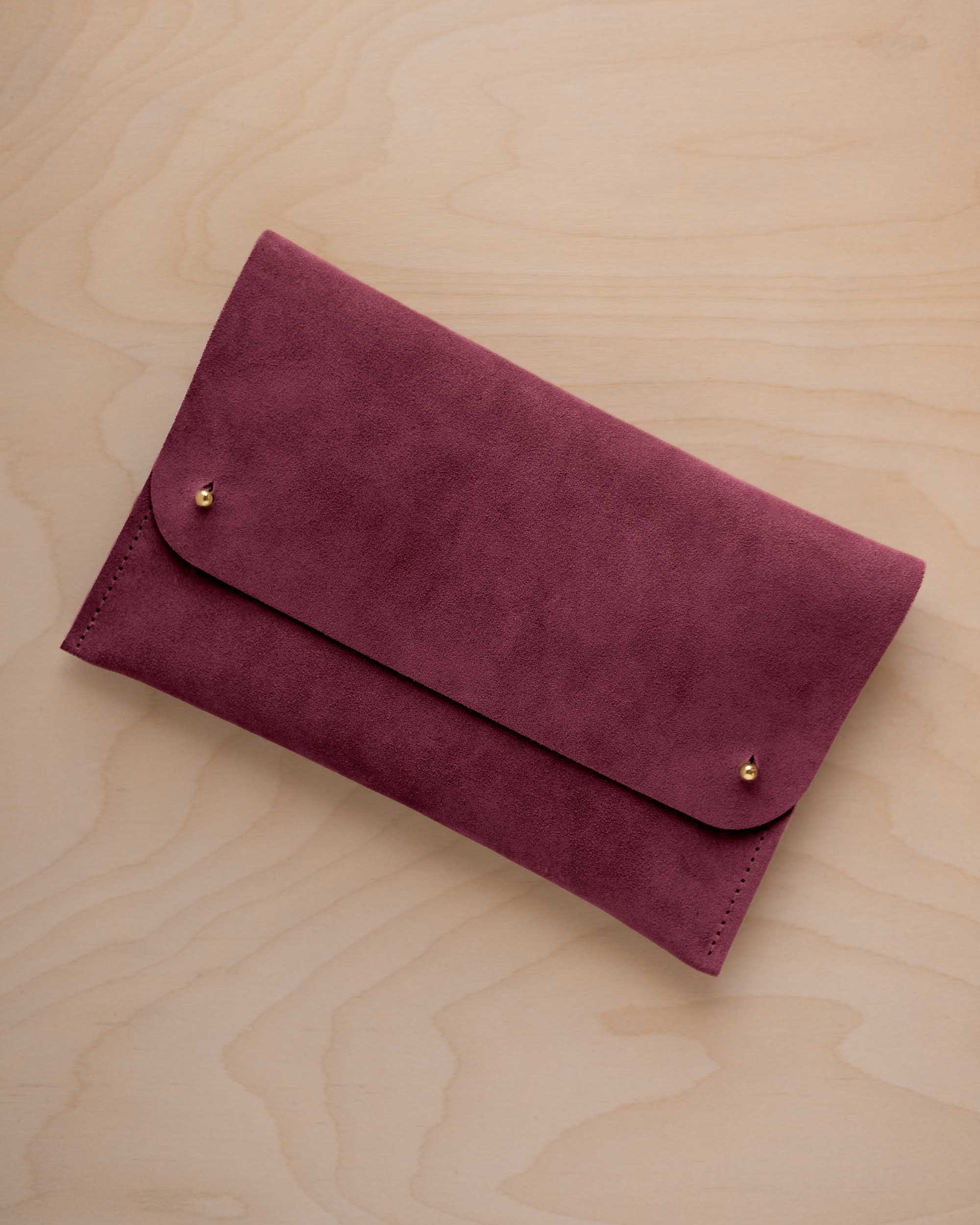A burgundy suede clutch with small gold coloured metal studs, displayed diagonally on a light wood grain surface.