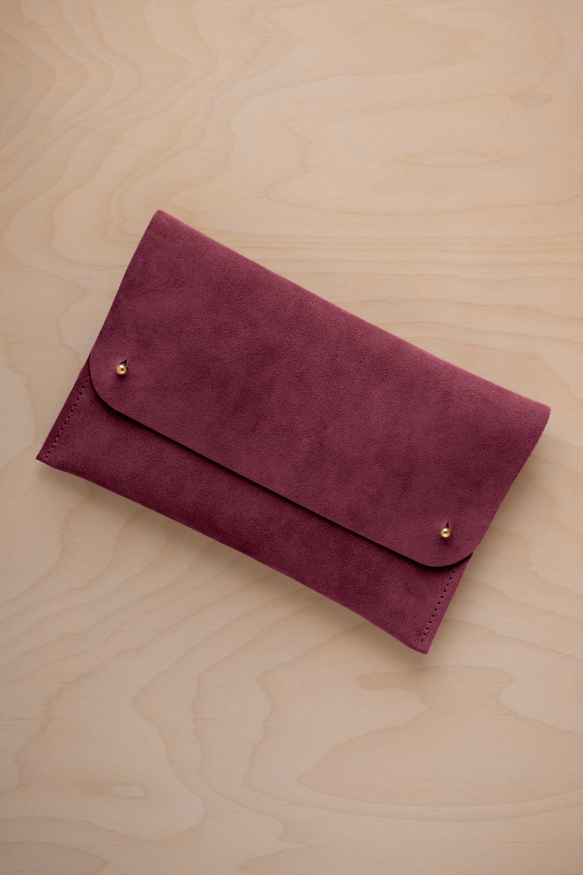 A burgundy suede clutch with small gold coloured metal studs, displayed diagonally on a light wood grain surface.