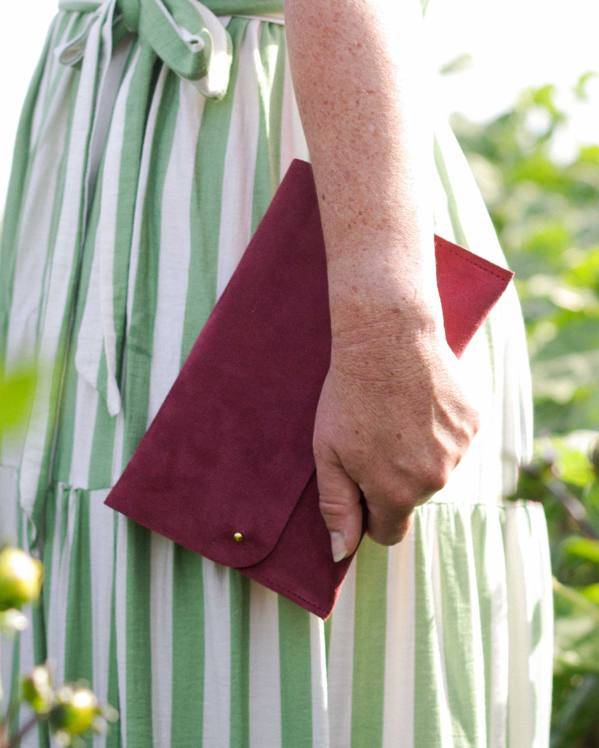 A close-up of a person wearing a green and white striped dress, holding a burgundy suede envelope clutch with a gold snap button.