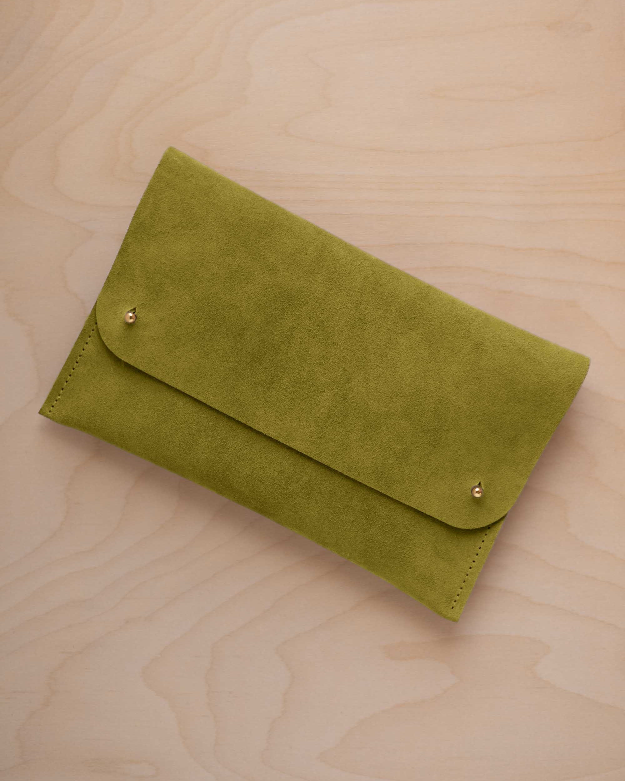 A chartreuse lime green coloured suede clutch with small gold coloured metal studs, displayed diagonally on a light wood grain surface.