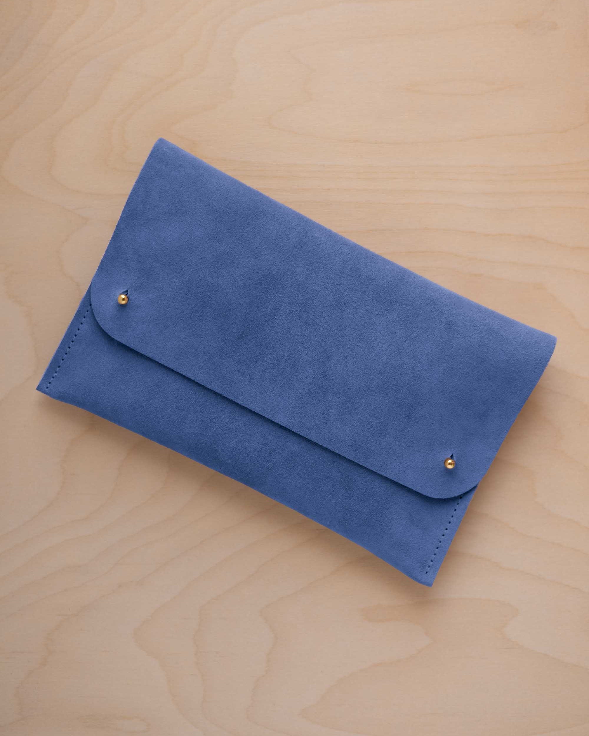 A cobalt blue suede clutch with small gold coloured metal studs, displayed diagonally on a light wood grain surface.