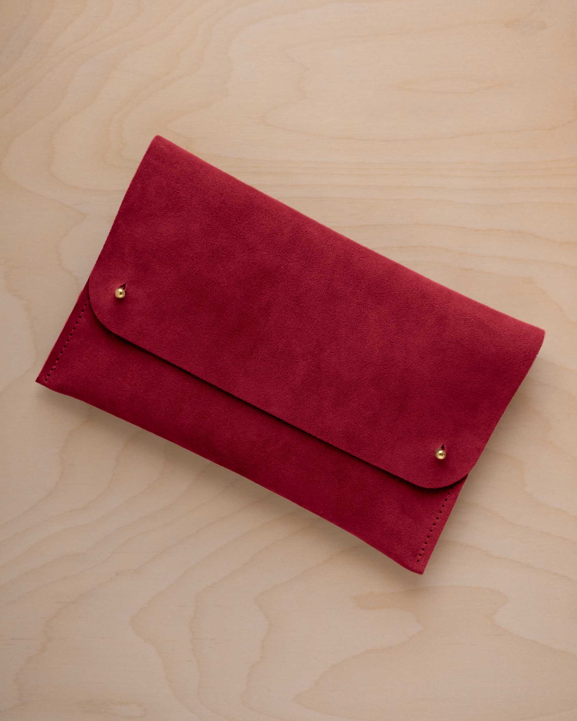 A crimson red suede clutch with small gold coloured metal studs, displayed diagonally on a light wood grain surface.