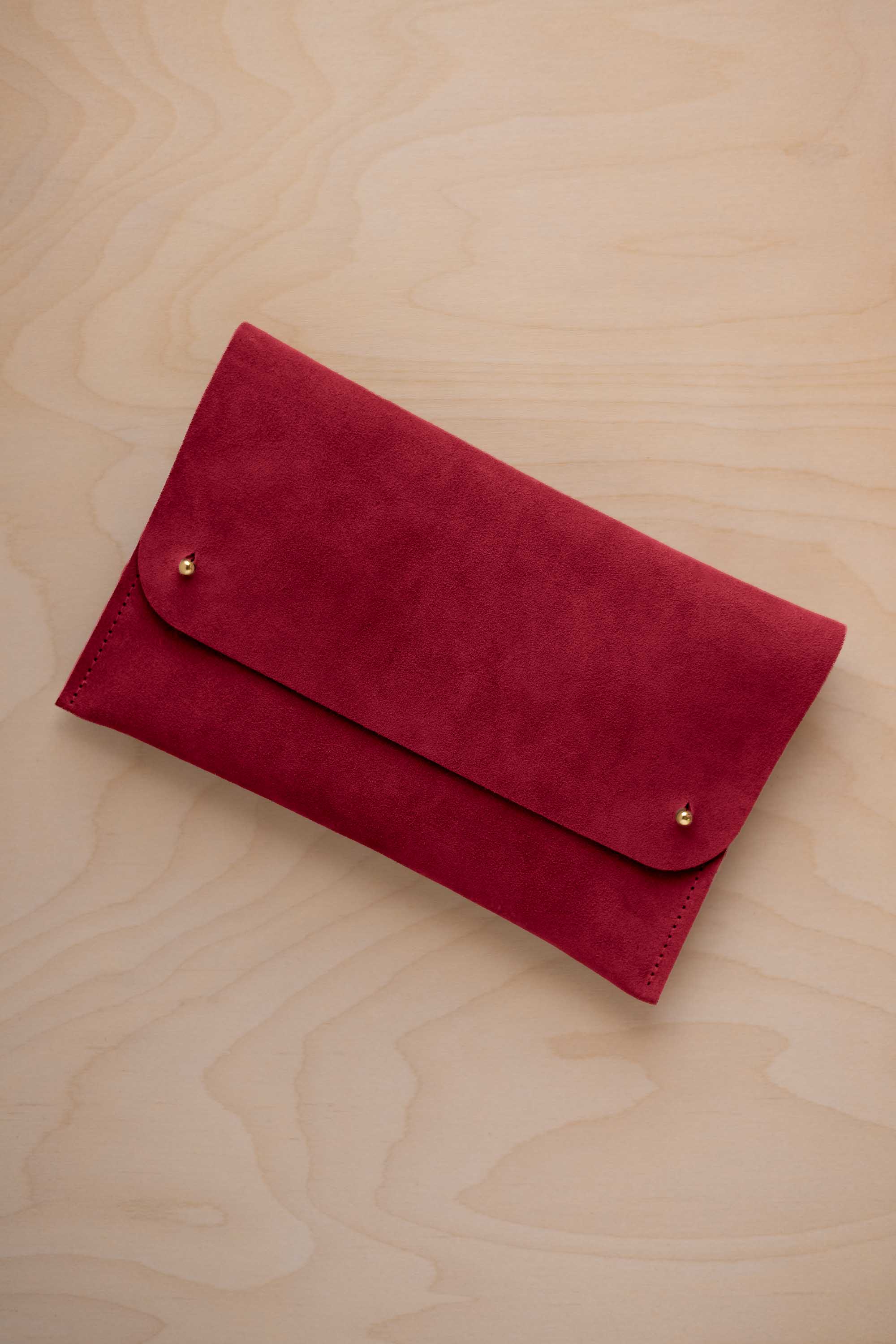 A crimson red suede clutch with small gold coloured metal studs, displayed diagonally on a light wood grain surface.