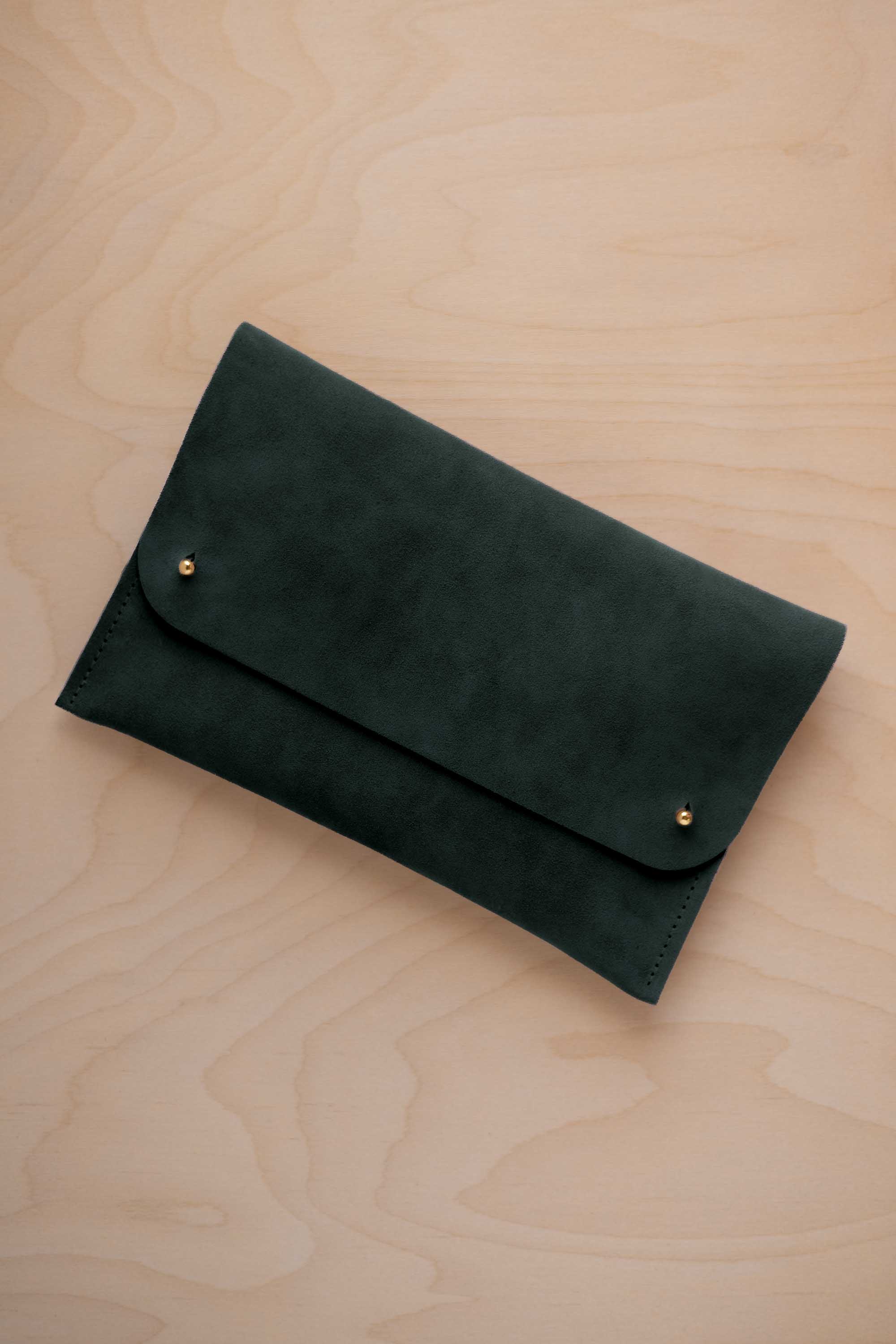 A deep forest green coloured suede clutch with small gold coloured metal studs, displayed diagonally on a light wood grain surface.