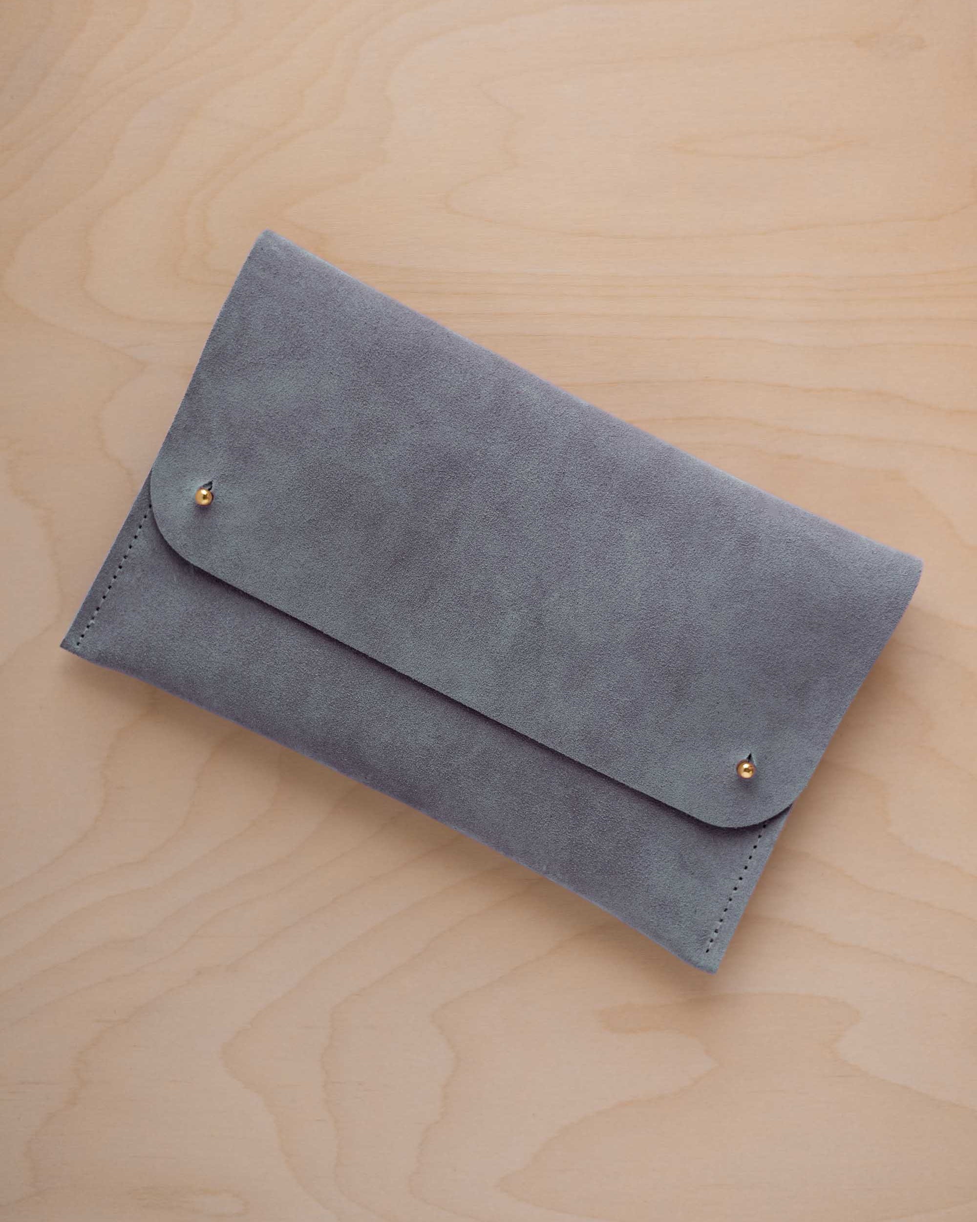 A dusky blue suede clutch with small gold coloured metal studs, displayed diagonally on a light wood grain surface.