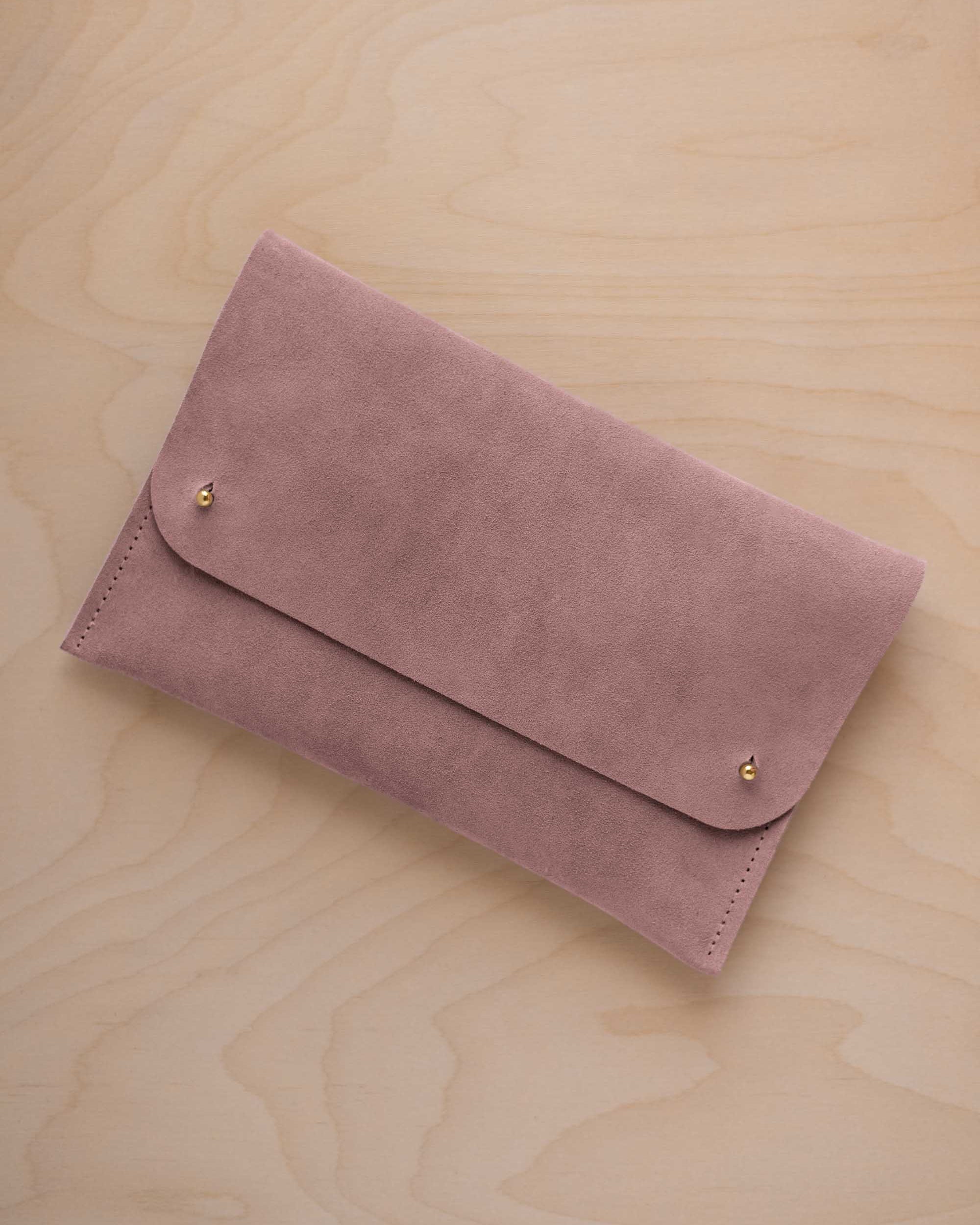 A dusty mauve grey suede clutch with small gold coloured metal studs, displayed diagonally on a light wood grain surface.