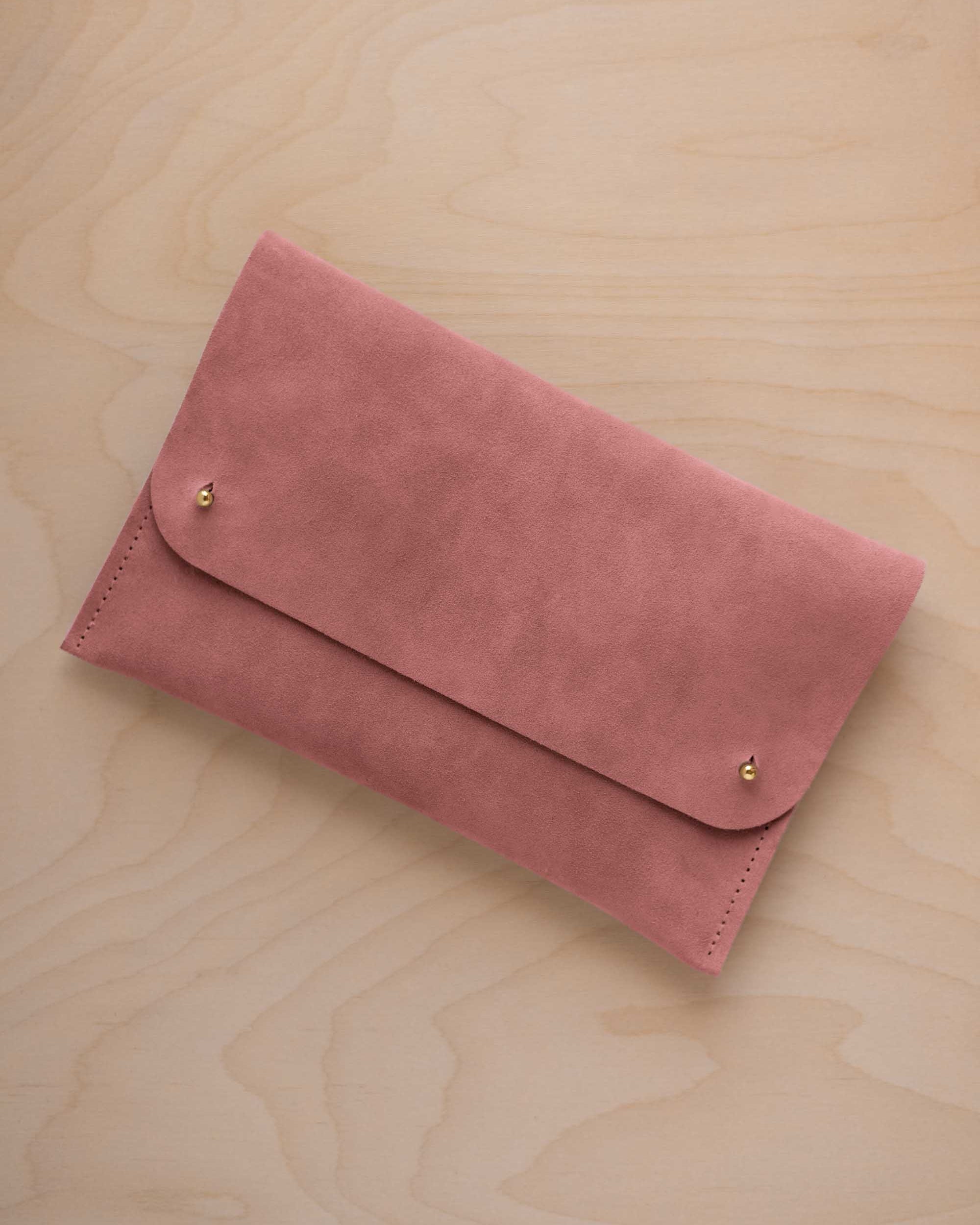 A dusty rose pink coloured suede clutch with small gold coloured metal studs, displayed diagonally on a light wood grain surface.