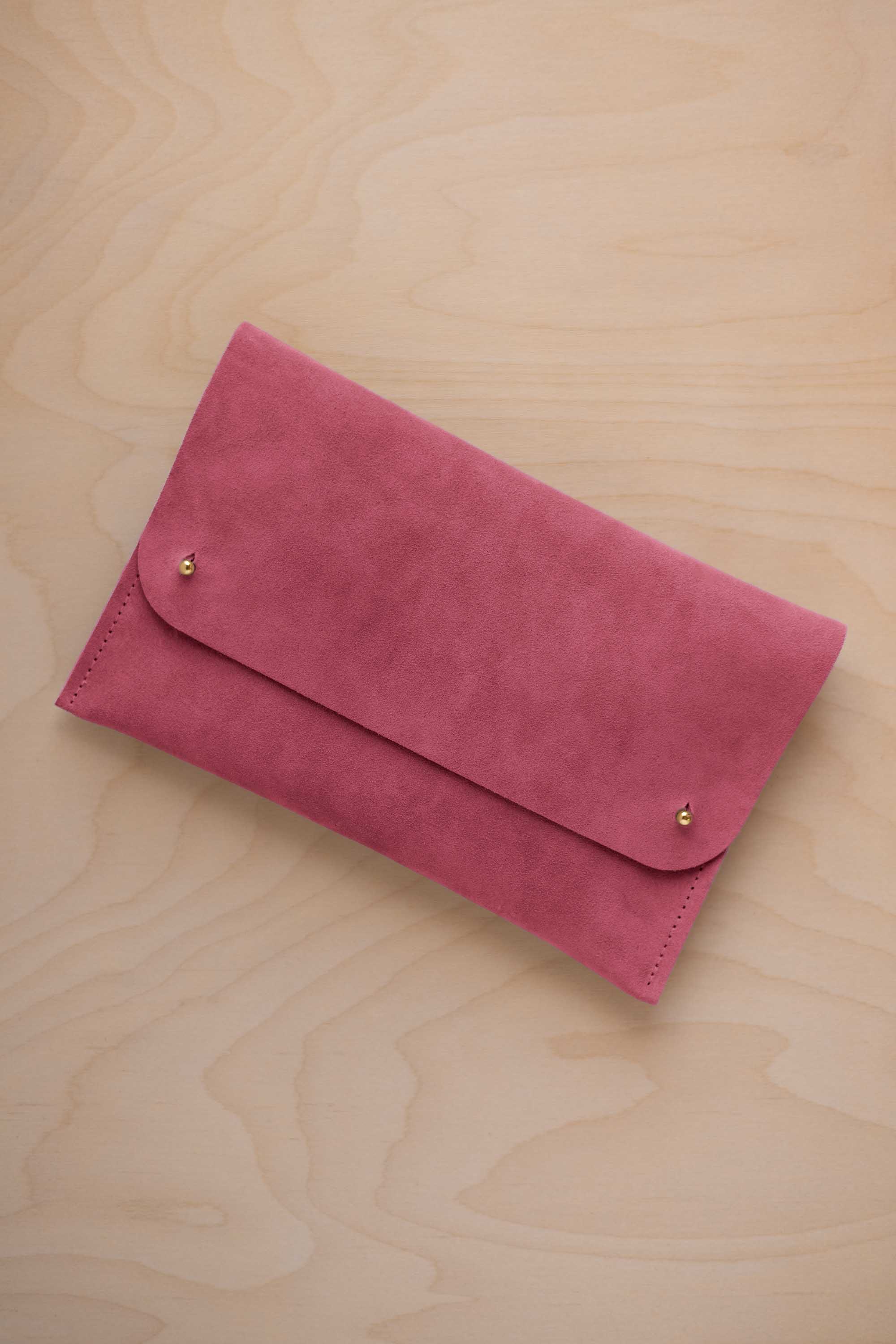 A faded rose pink suede clutch with small gold coloured metal studs, displayed diagonally on a light wood grain surface.