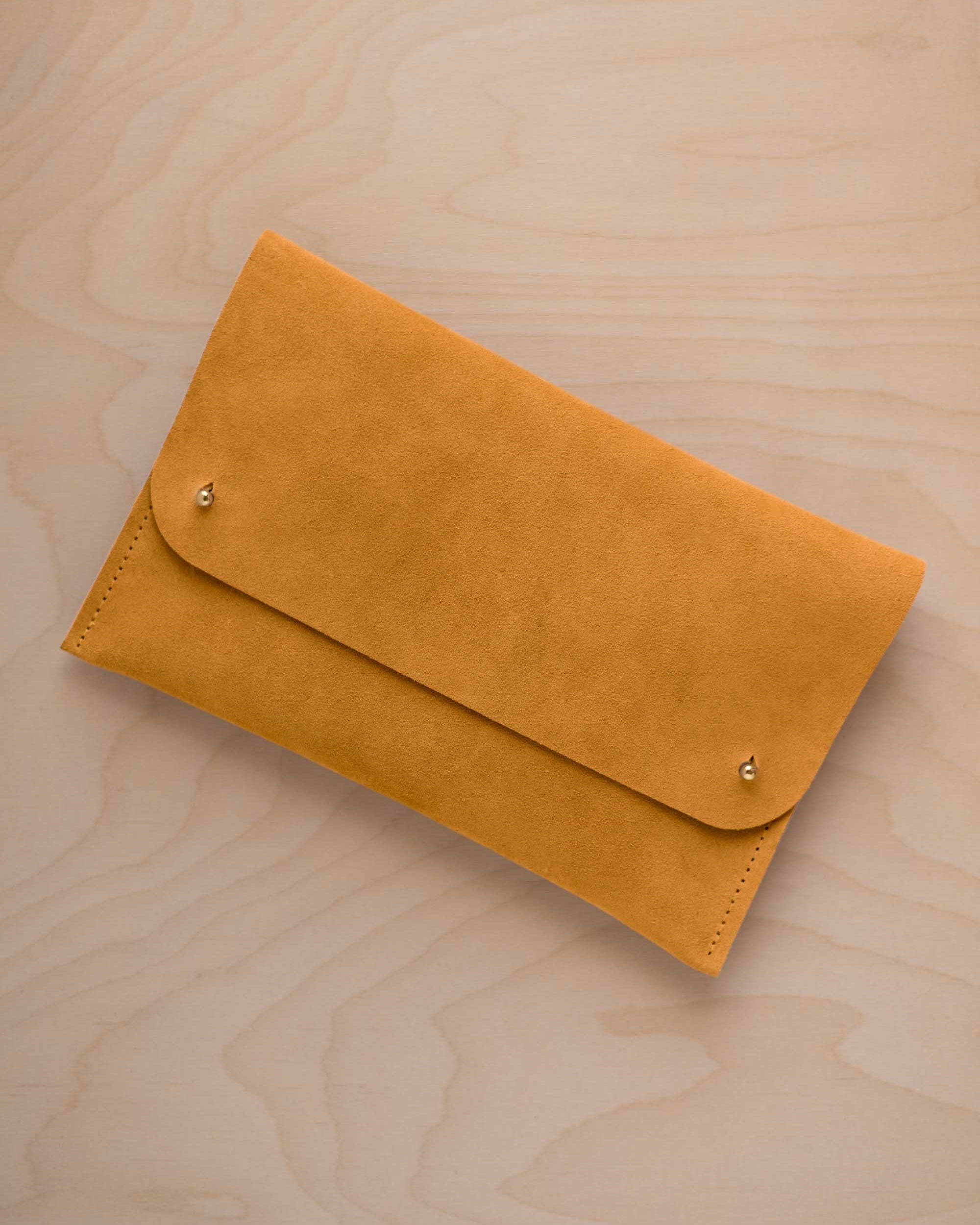 A golden ochre mustard yellow coloured suede clutch with small gold coloured metal studs, displayed diagonally on a light wood grain surface.