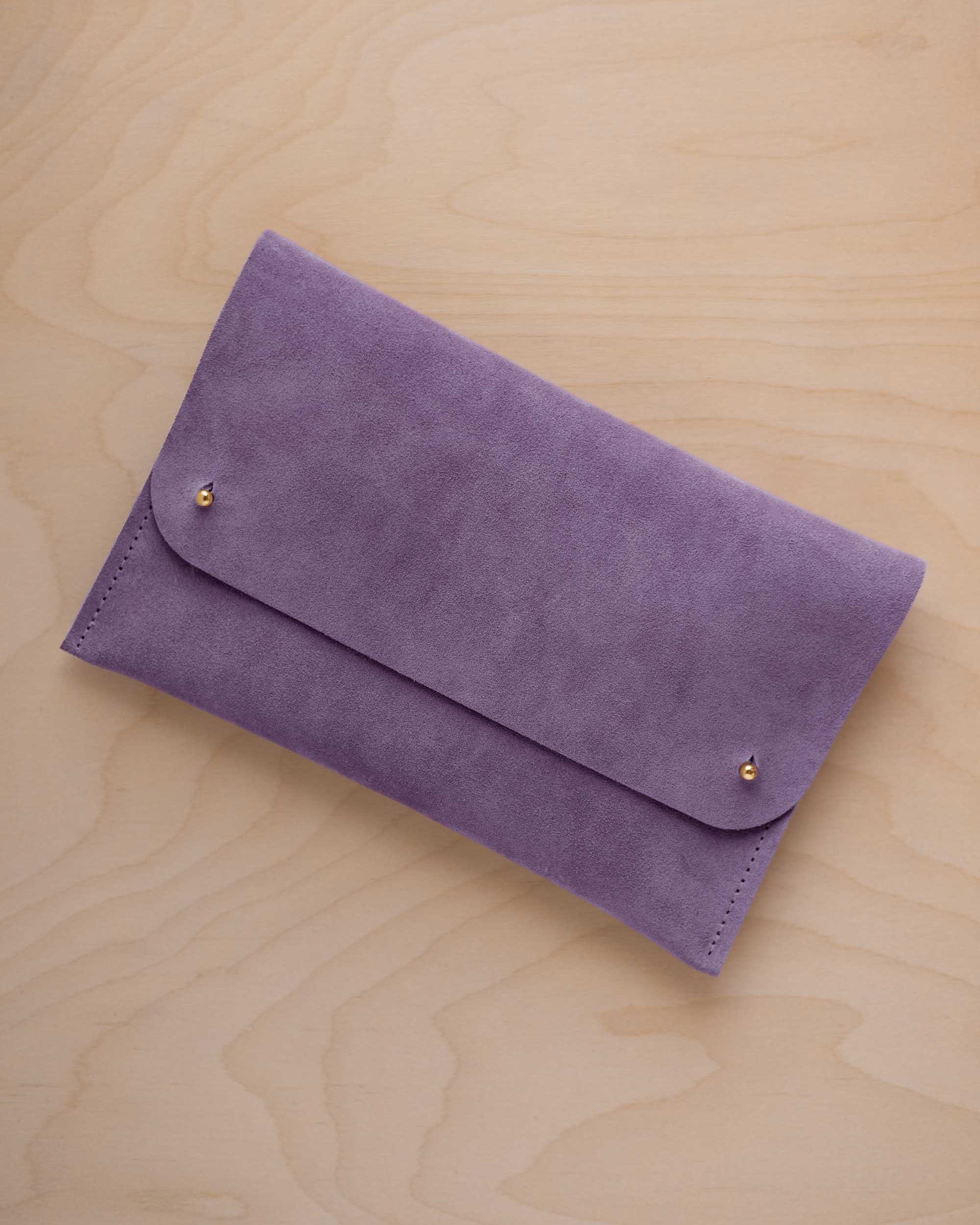 A lavender coloured suede clutch with small gold coloured metal studs, displayed diagonally on a light wood grain surface.