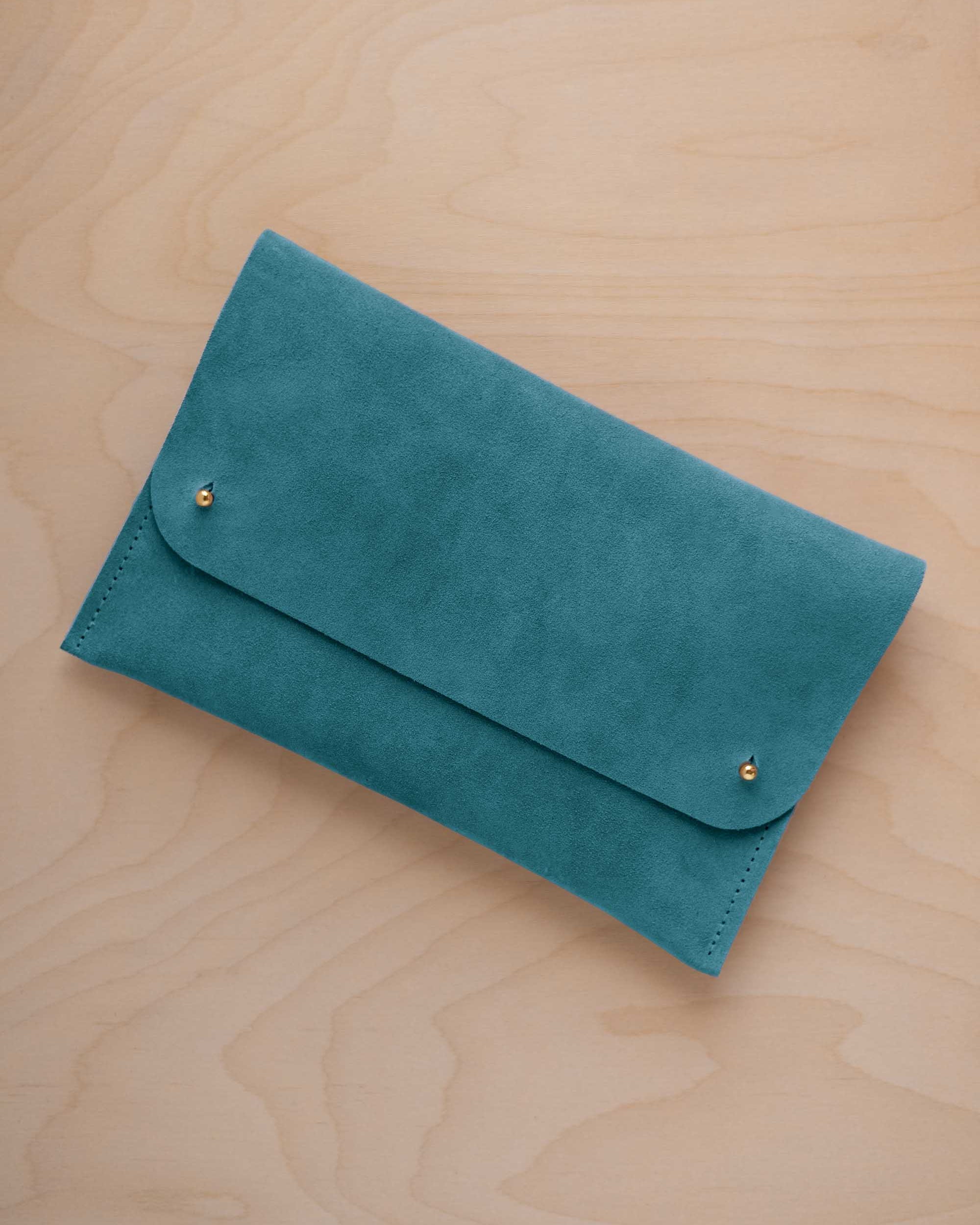 A marine teal coloured suede clutch with small gold coloured metal studs, displayed diagonally on a light wood grain surface.