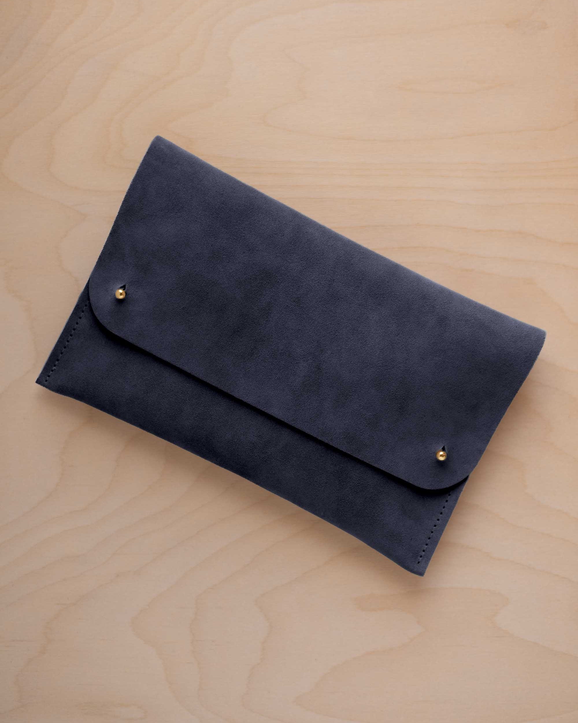 A midnight blue coloured suede clutch with small gold coloured metal studs, displayed diagonally on a light wood grain surface.