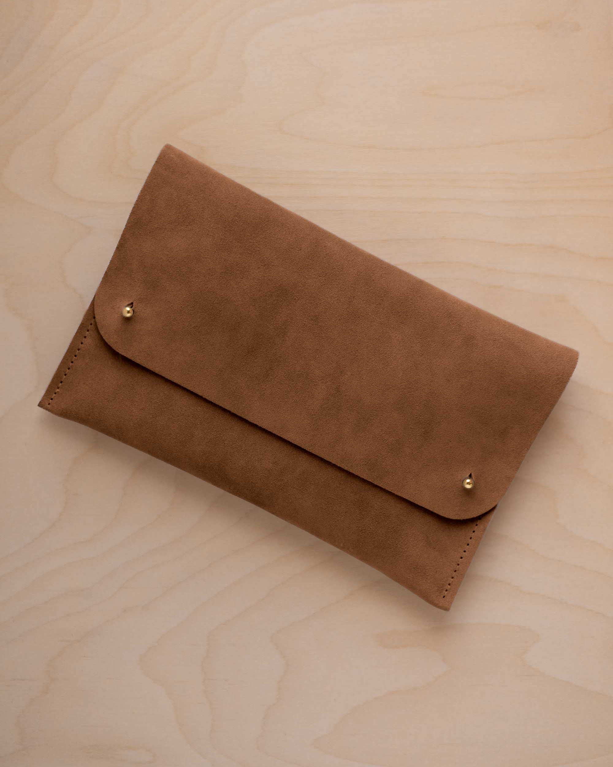 A mocha brown coloured suede clutch with small gold coloured metal studs, displayed diagonally on a light wood grain surface.