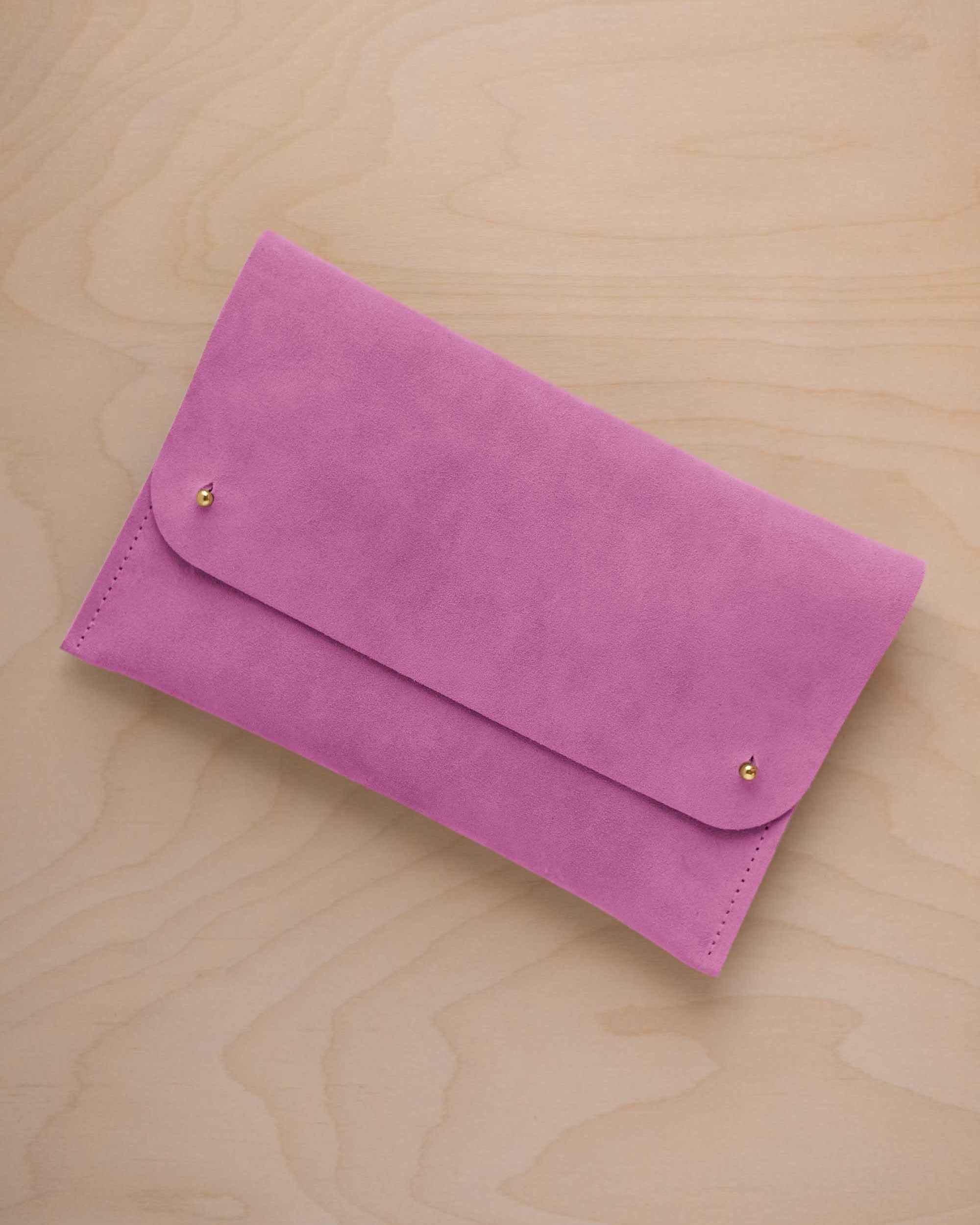 A peony pink suede clutch with small gold coloured metal studs, displayed diagonally on a light wood grain surface.