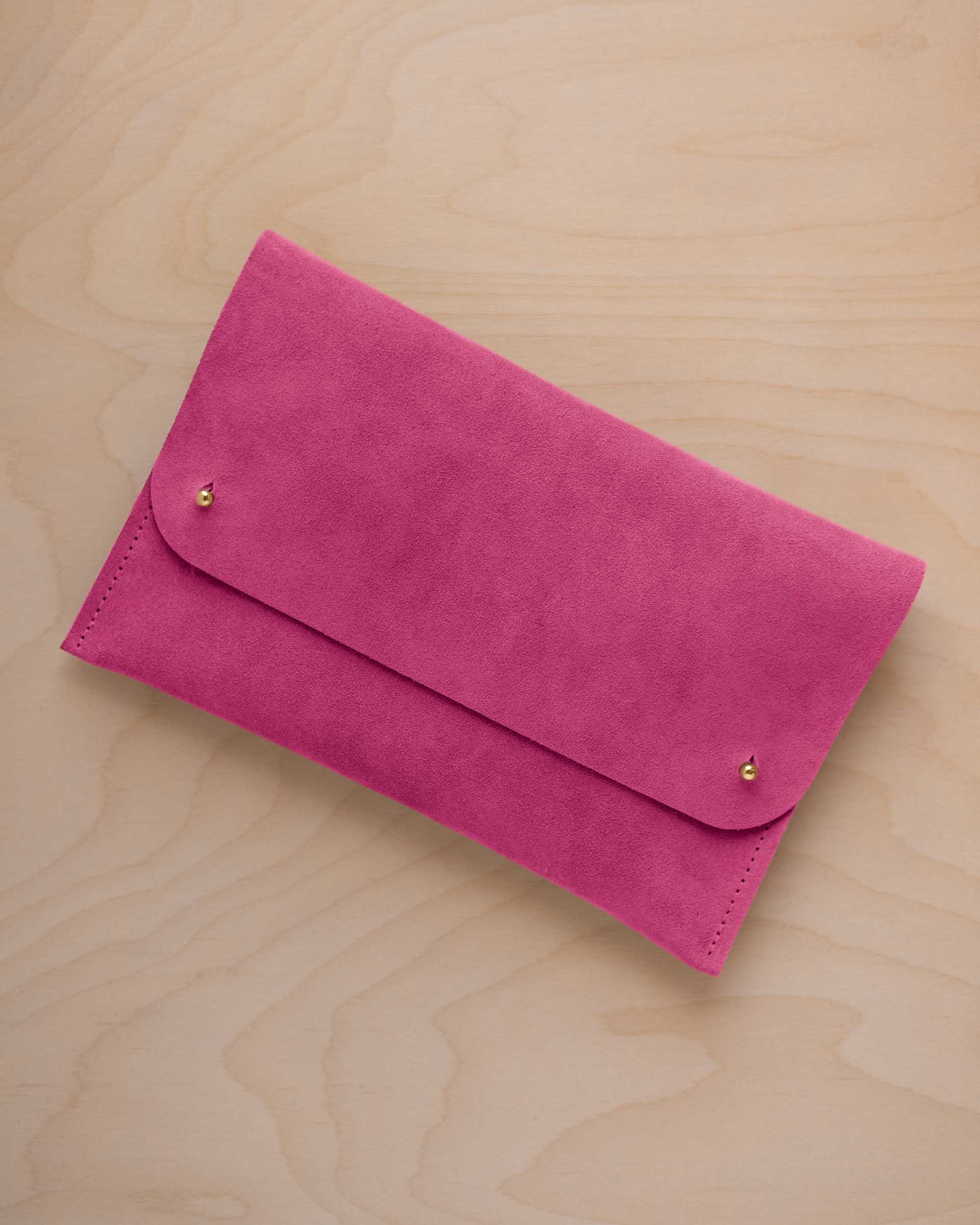 A raspberry pink coloured suede clutch with small gold coloured metal studs, displayed diagonally on a light wood grain surface.
