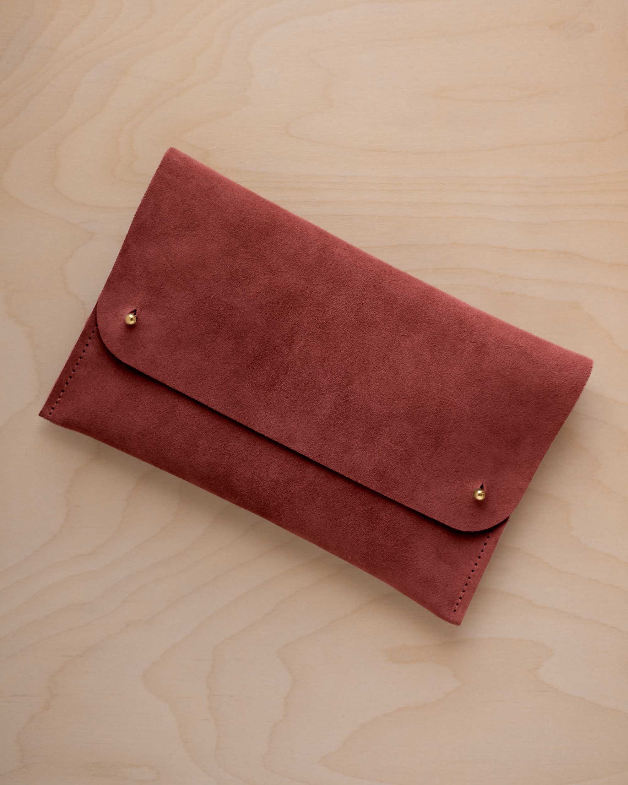 A rust red suede clutch with small gold coloured metal studs, displayed diagonally on a light wood grain surface.