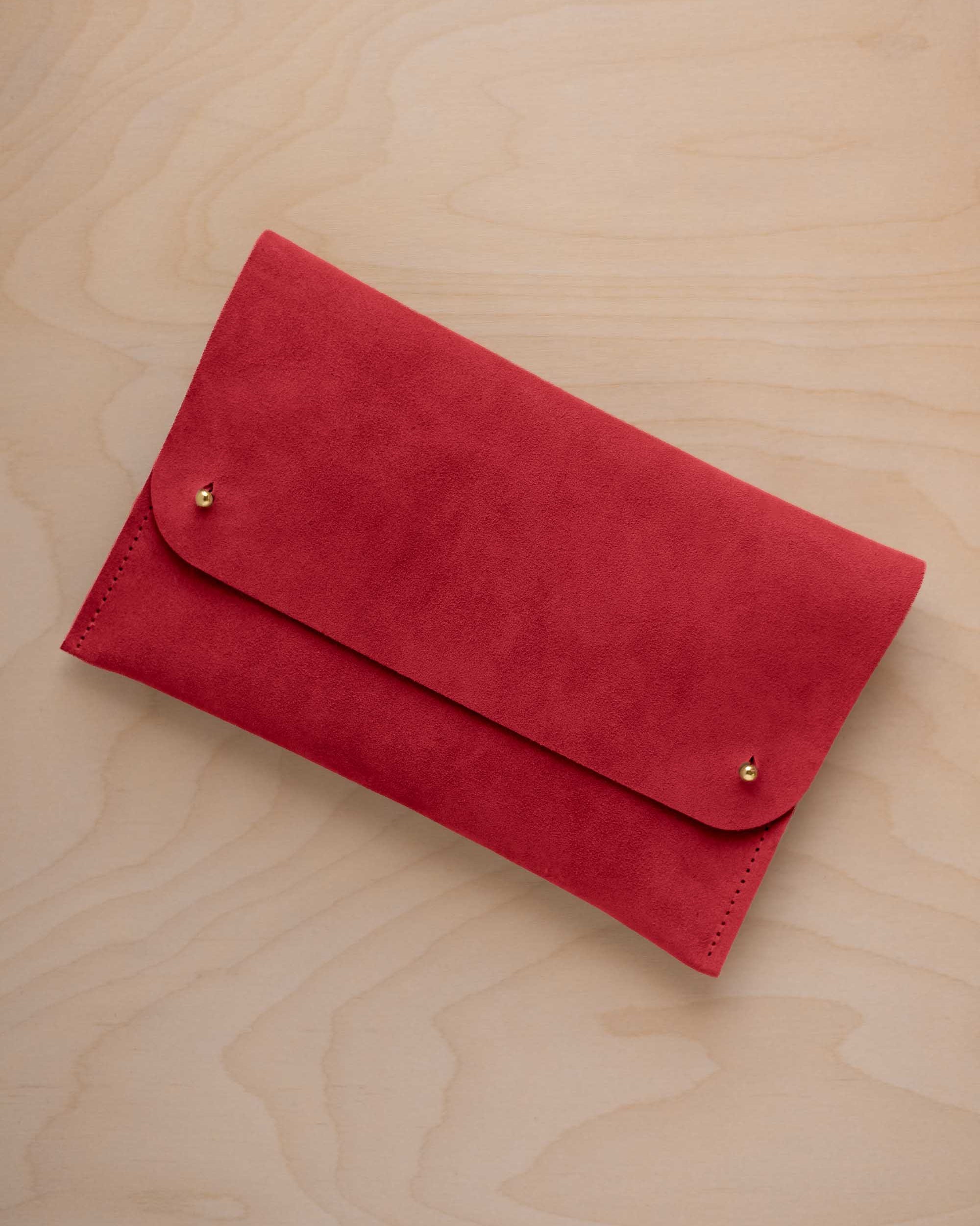 A scarlet red suede clutch with small gold coloured metal studs, displayed diagonally on a light wood grain surface.