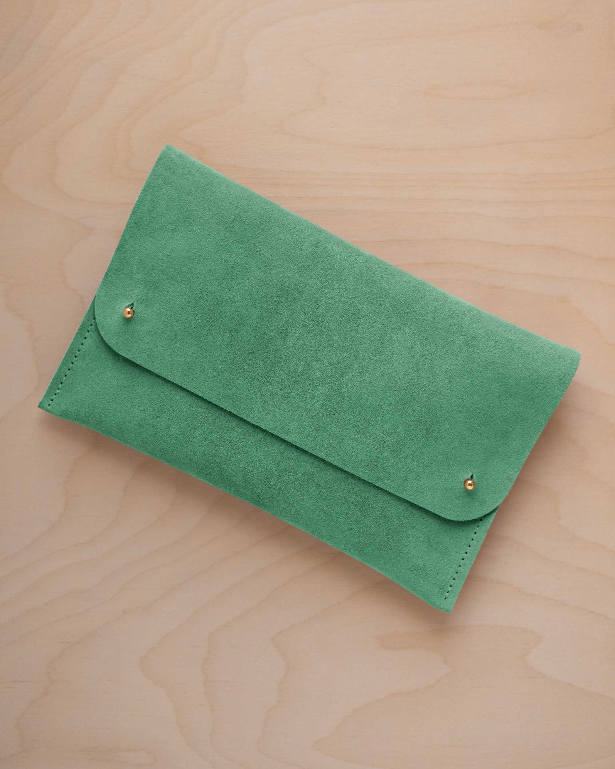 A seafoam sage green coloured suede clutch with small gold coloured metal studs, displayed diagonally on a light wood grain surface.