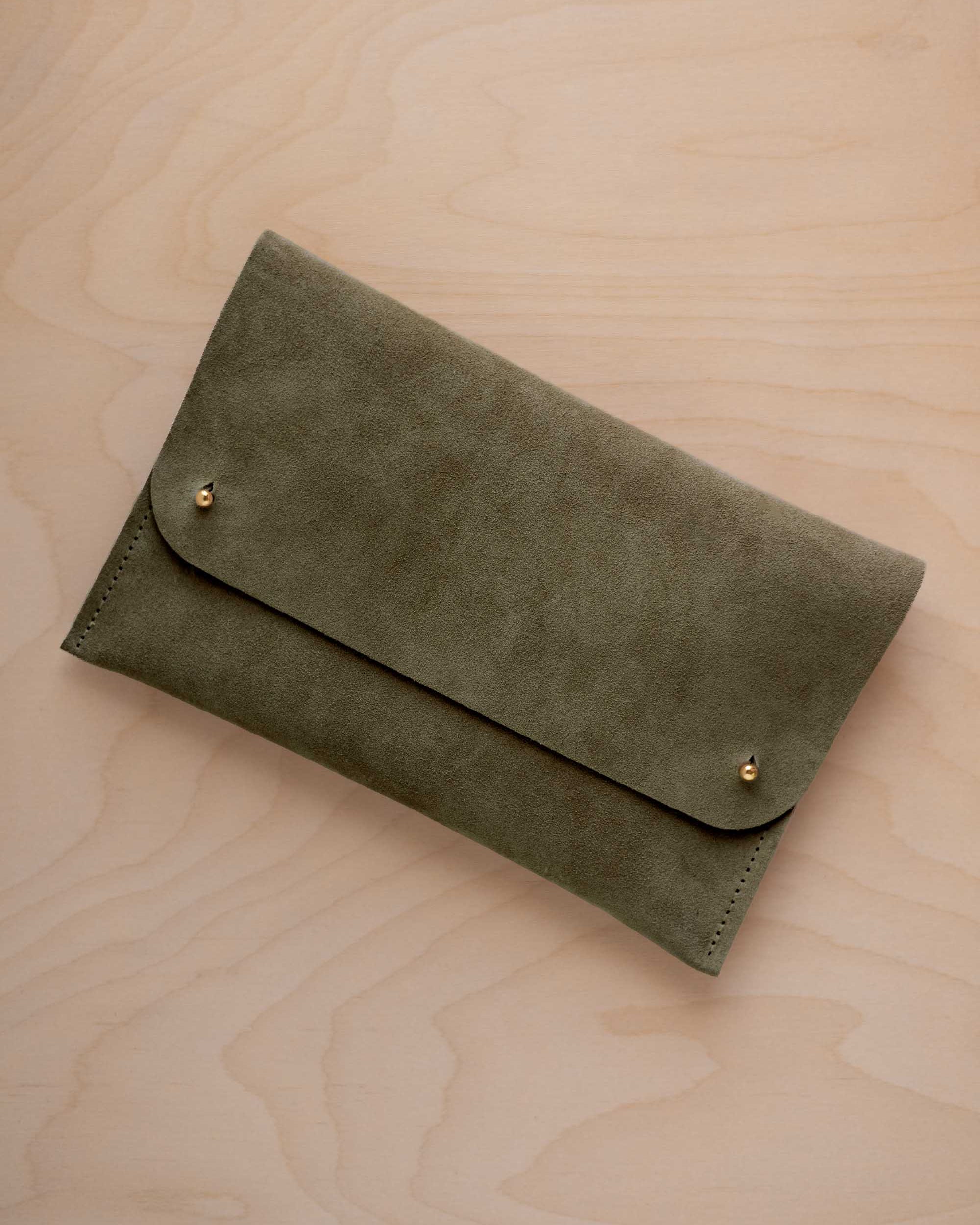 An olive green coloured suede clutch with small gold coloured metal studs, displayed diagonally on a light wood grain surface.