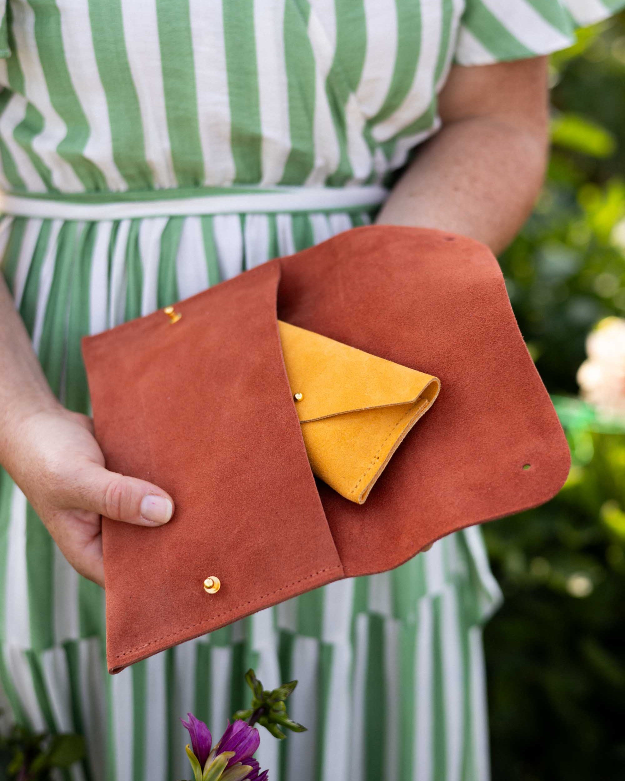 A person in a green and white striped dress holds an open terracotta brown suede pouch or clutch bag, showing a small yellow envelope-style purse inside. The background is a softly blurred green outdoor setting with a purple flower in the foreground.
