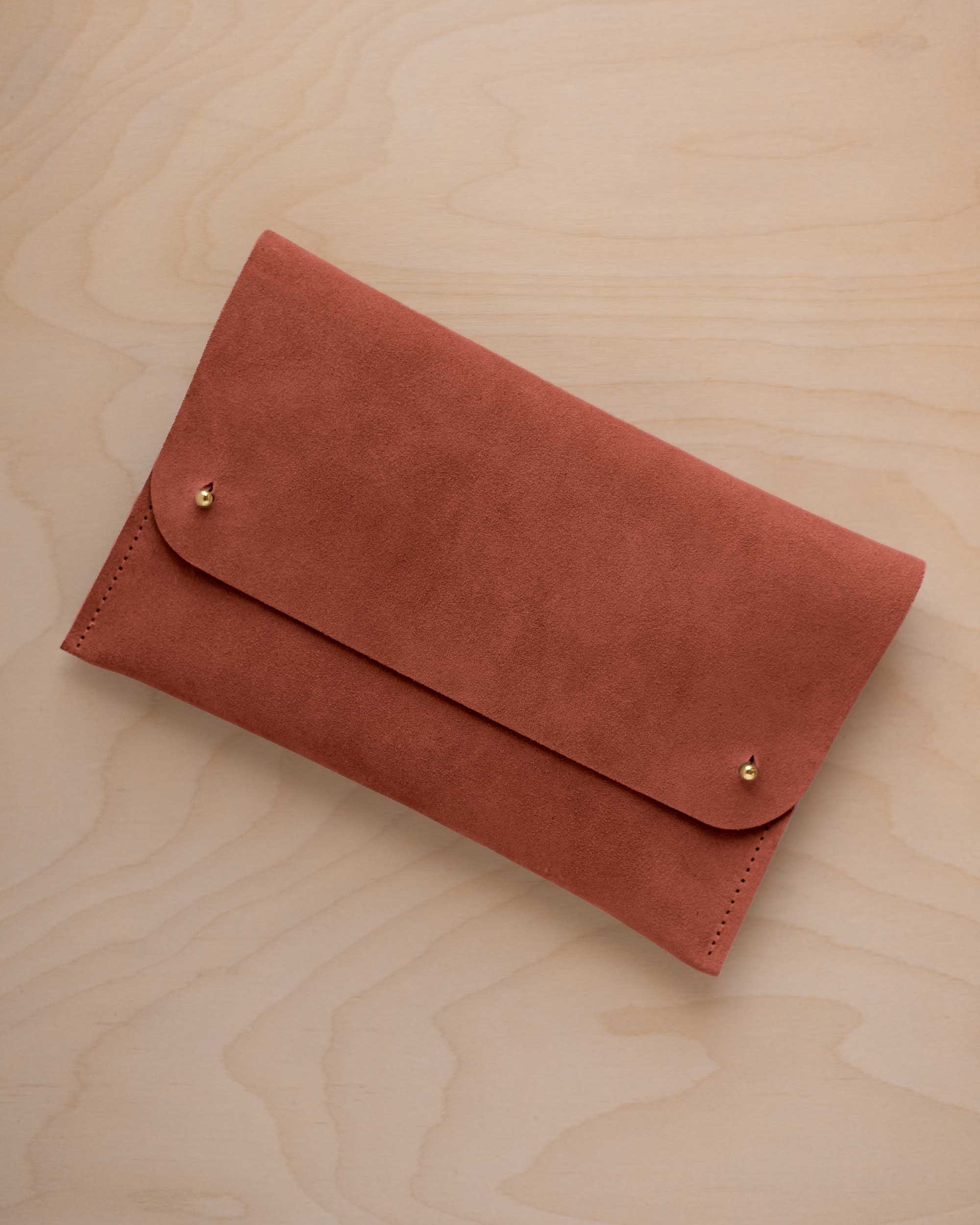 A terracotta coloured suede clutch with small gold coloured metal studs, displayed diagonally on a light wood grain surface.