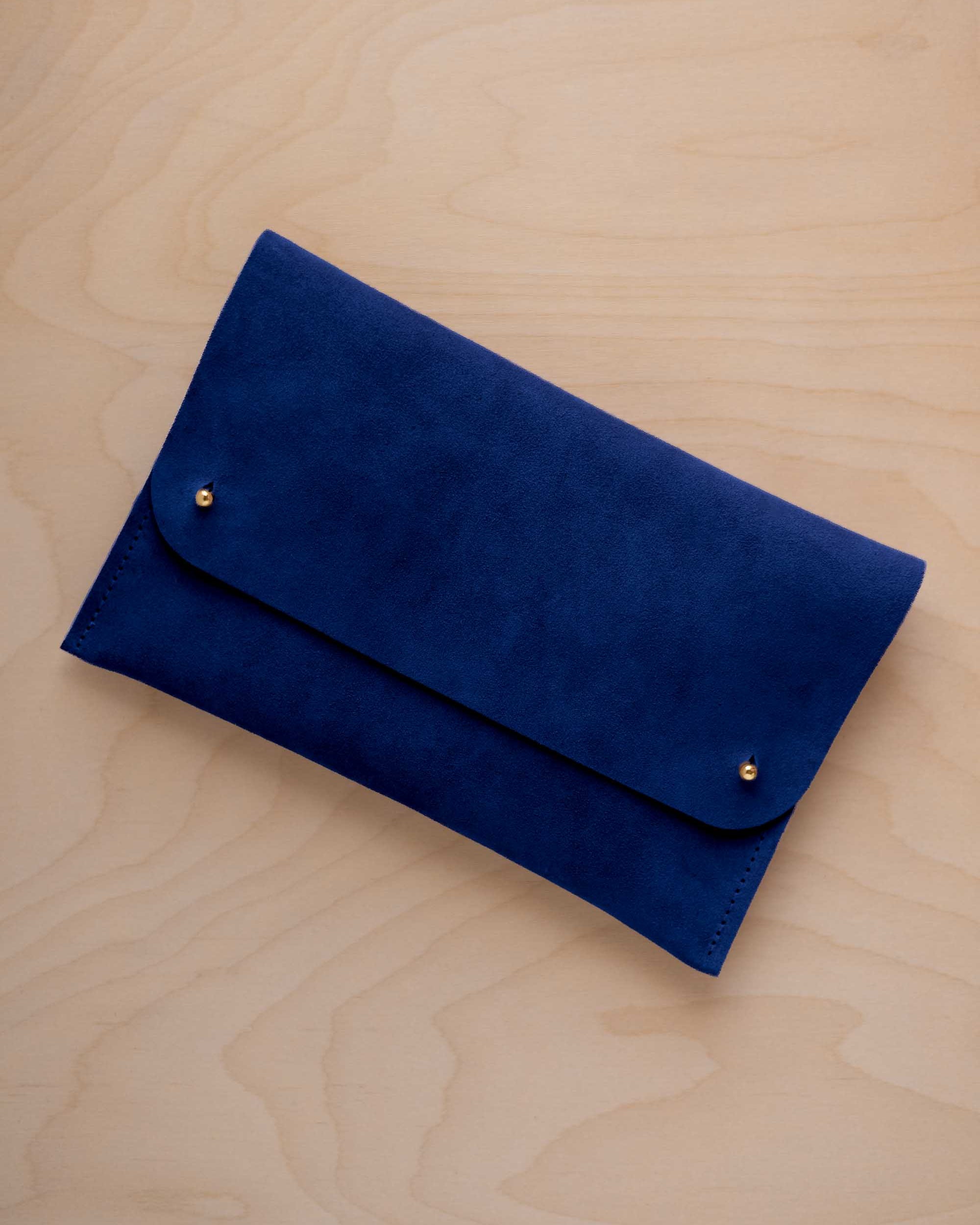 An ultramarine blue coloured suede clutch with small gold coloured metal studs, displayed diagonally on a light wood grain surface.