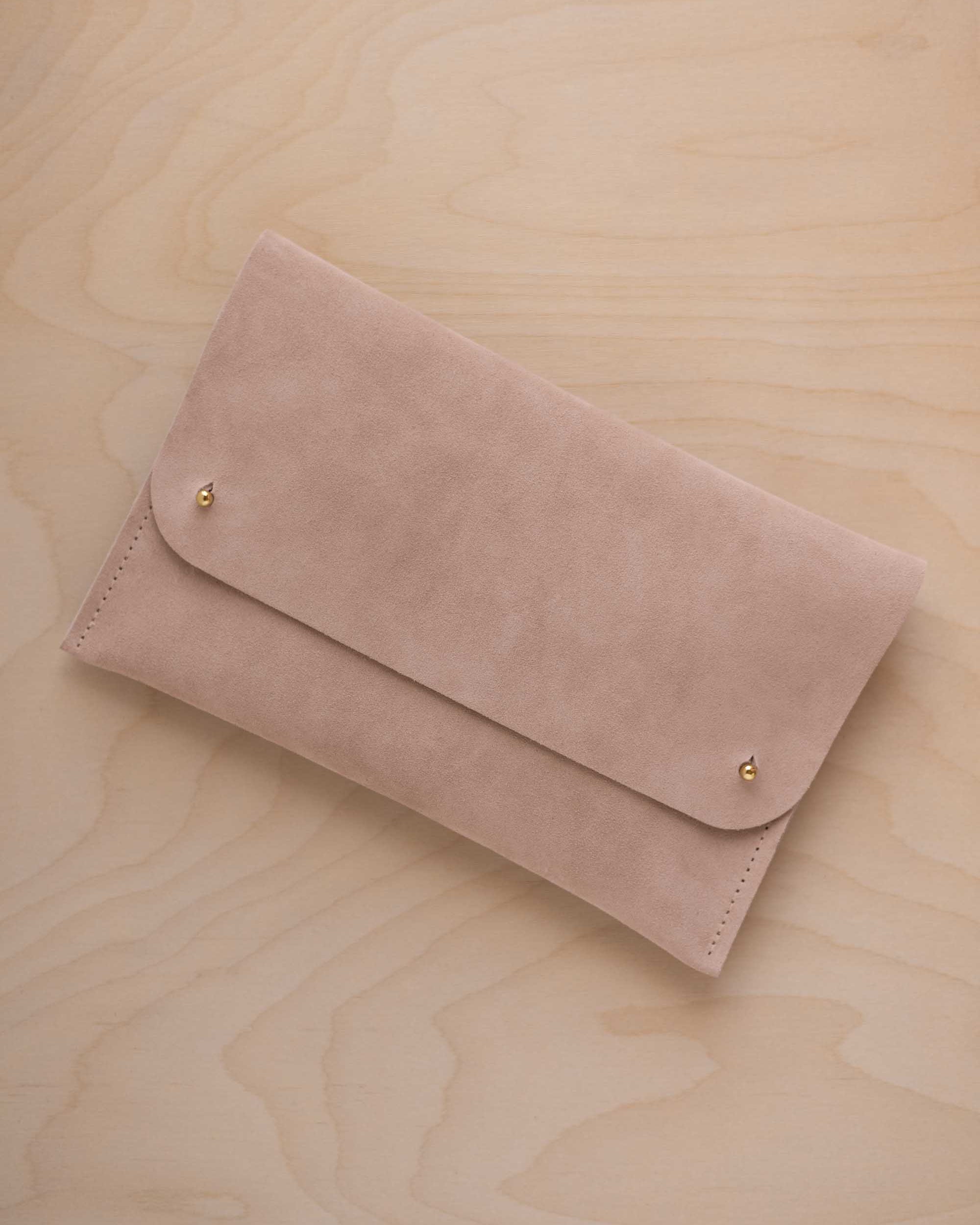 A warm beige suede clutch with small gold coloured metal studs, displayed diagonally on a light wood grain surface.