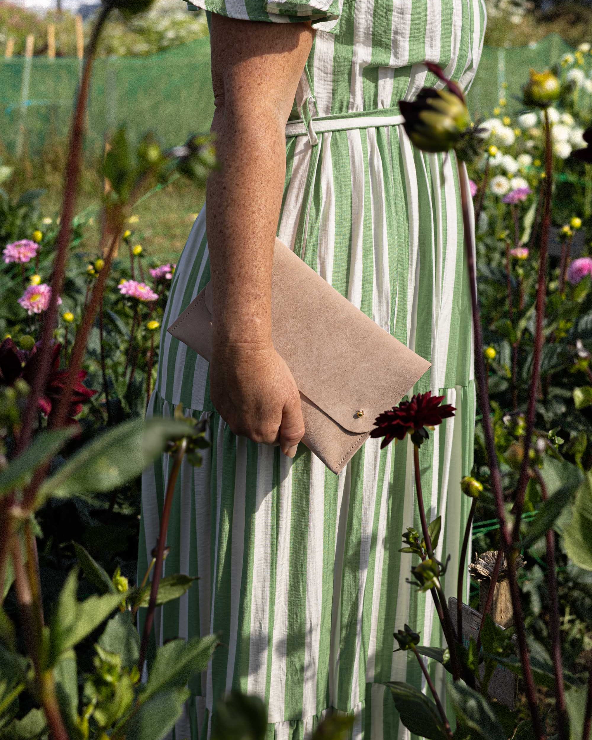 A person standing in a flower garden wearing a green and white vertically striped midi dress and holding a beige suede clutch bag.