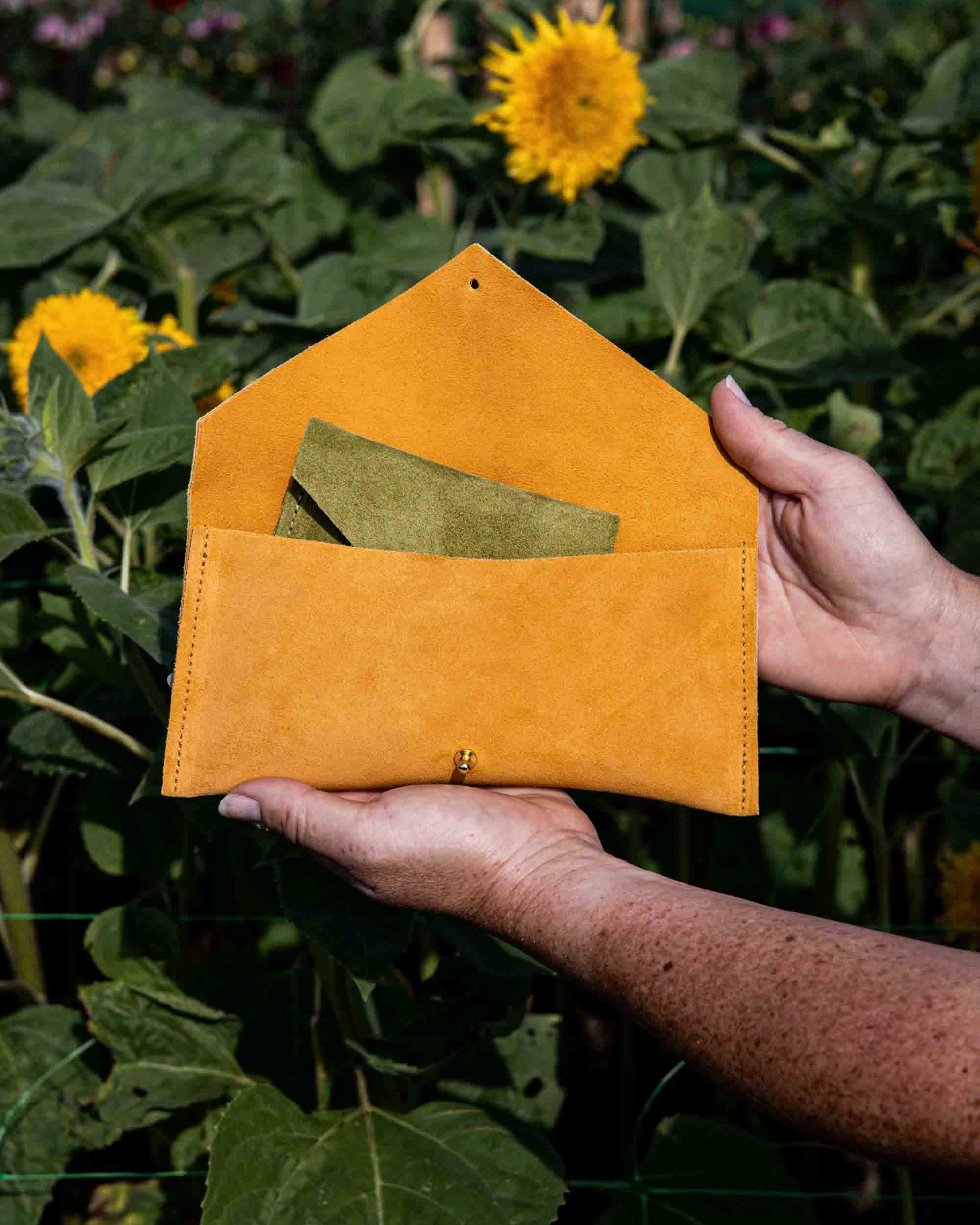 A mustard yellow-orange suede envelope-style pouch with a flap closure is held open by two hands in a sunflower field. A green suede purse is tucked inside the pouch.