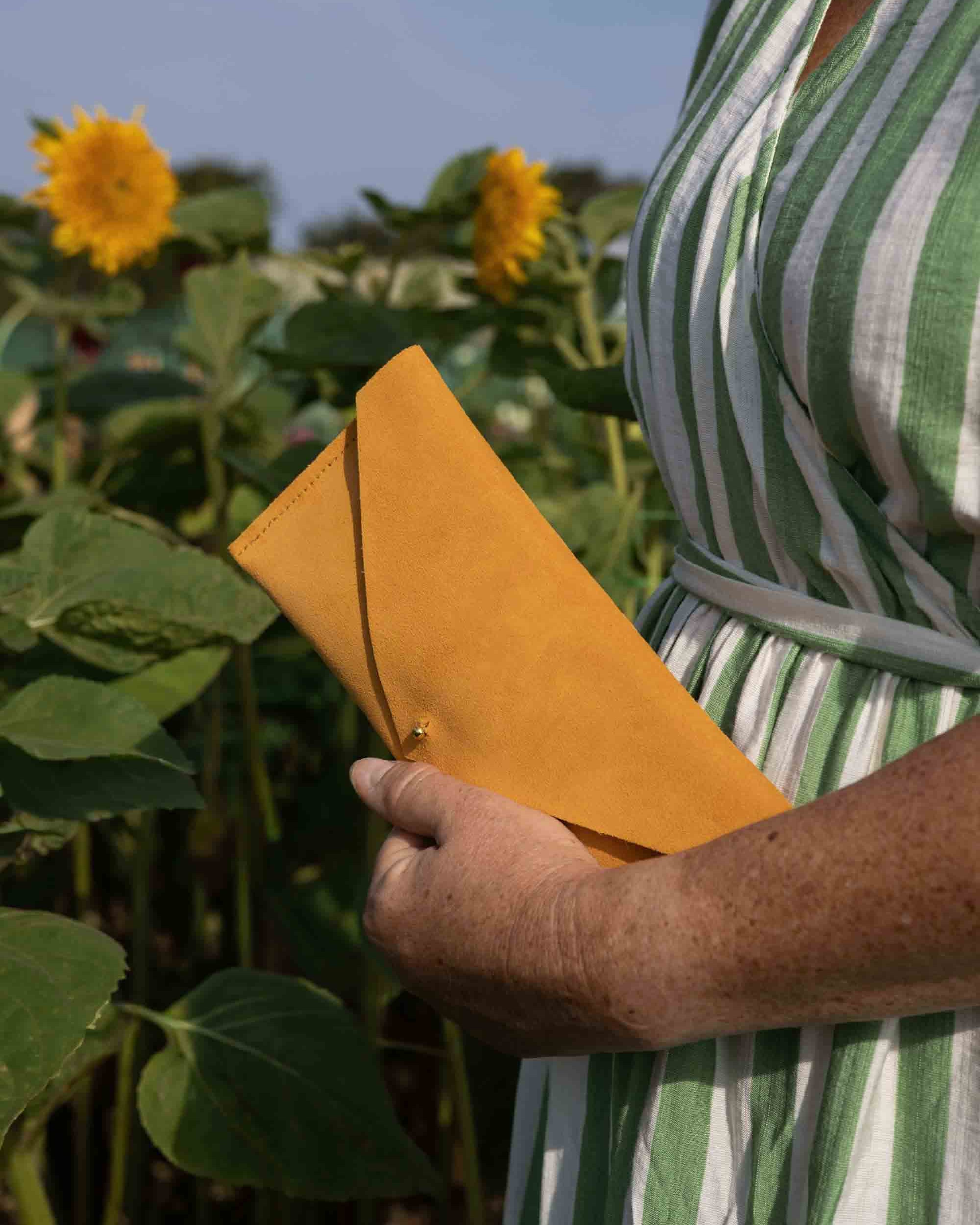 A person in a green and white striped dress holding a bright yellow-orange envelope-style clutch in a sunflower field.