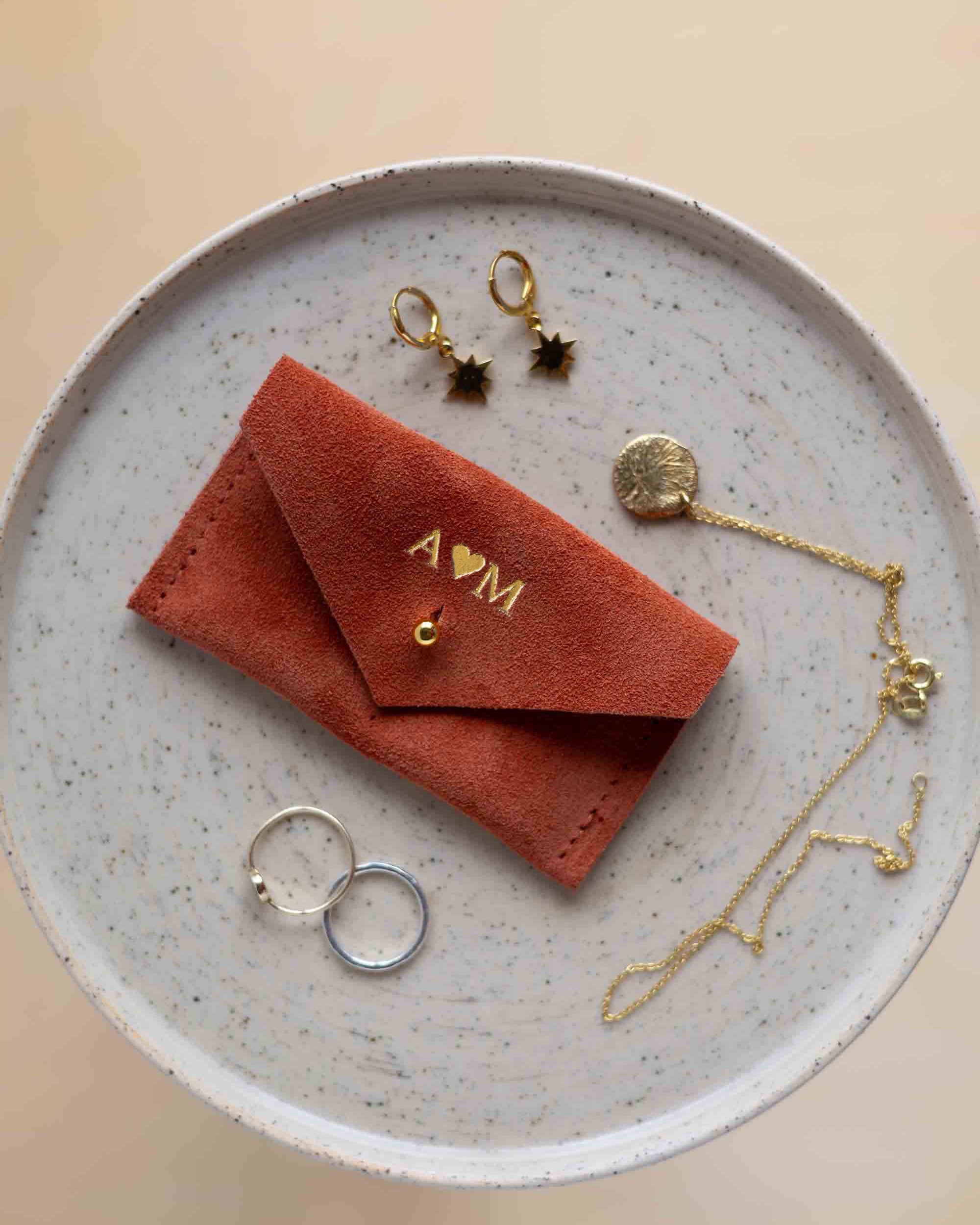 A top-down view of a terracotta coloured suede jewellery pouch with gold foiled initials with a gold coloured fastening, displayed on a round speckled ceramic plate alongside gold and silver jewellery pieces on a warm beige background.