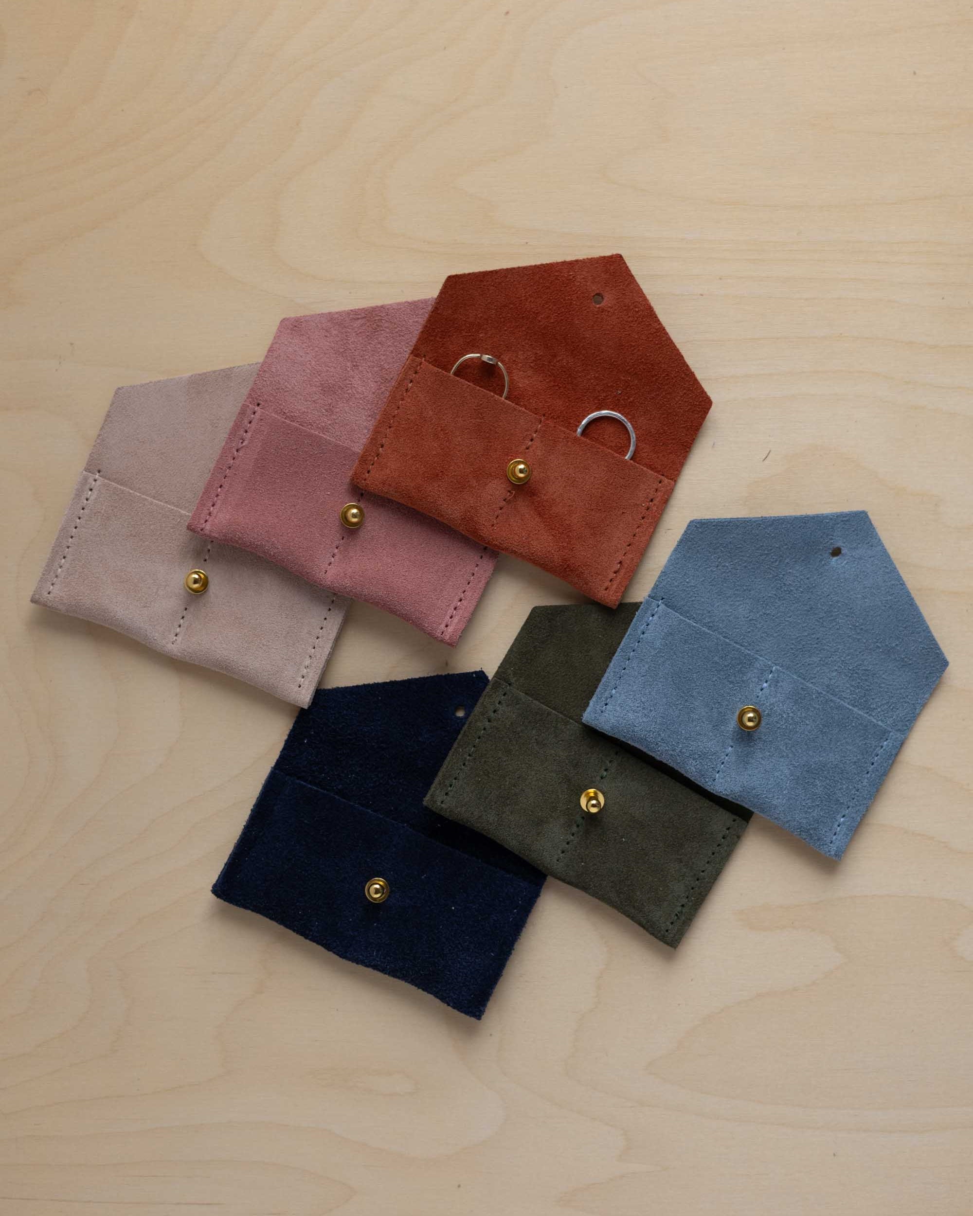 Six small suede pouches in assorted colours arranged together on a light wood background. The rust coloured suede pouch has couples wedding rings nestled inside.
