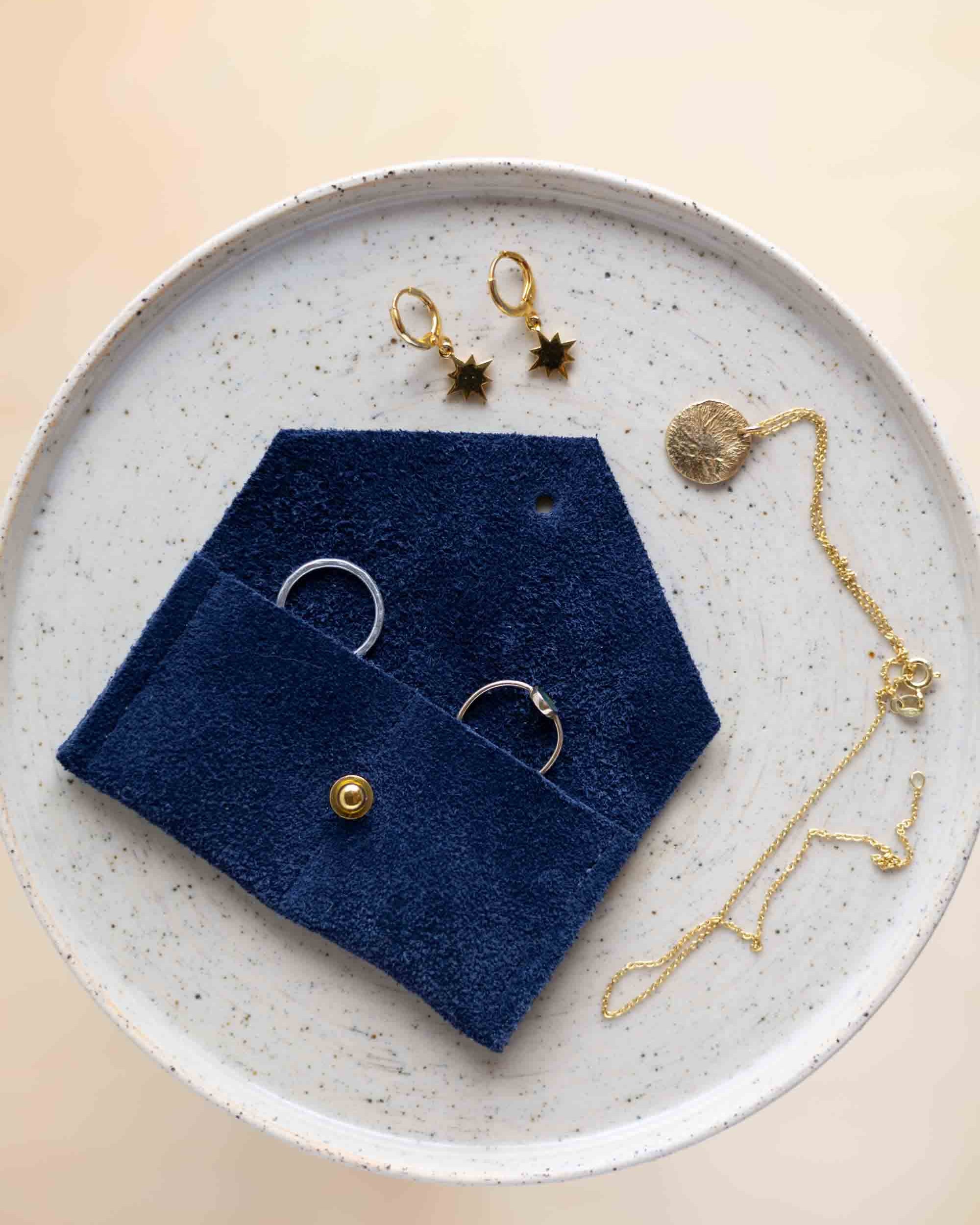 A top-down view of an ultramarine blue coloured suede jewellery pouch with a gold coloured fastening, displayed on a round speckled ceramic plate alongside gold and silver jewellery pieces on a warm beige background.