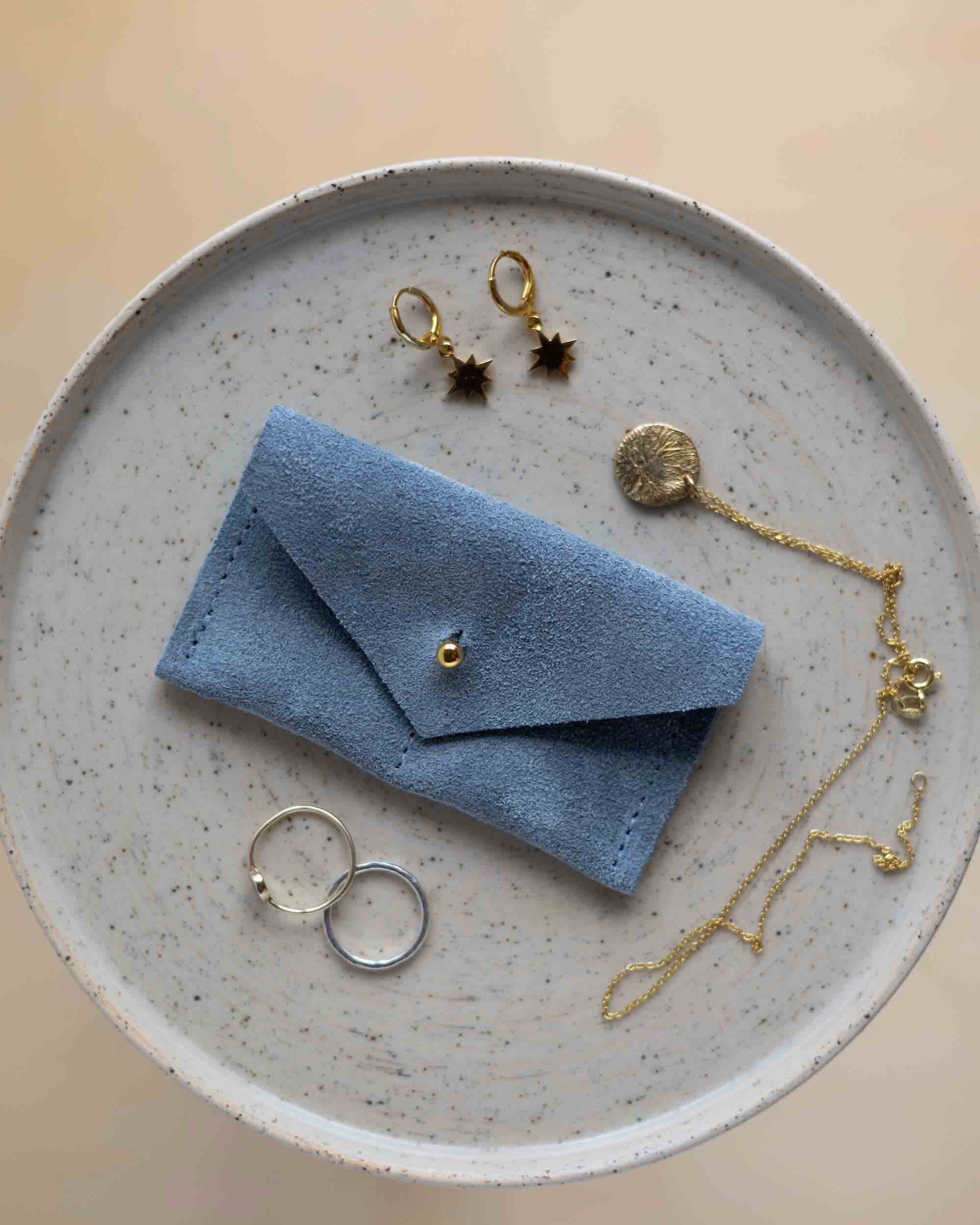 A top-down view of a dusky blue suede jewellery pouch with a gold coloured fastening, displayed on a round speckled ceramic plate alongside gold and silver jewellery pieces on a warm beige background.