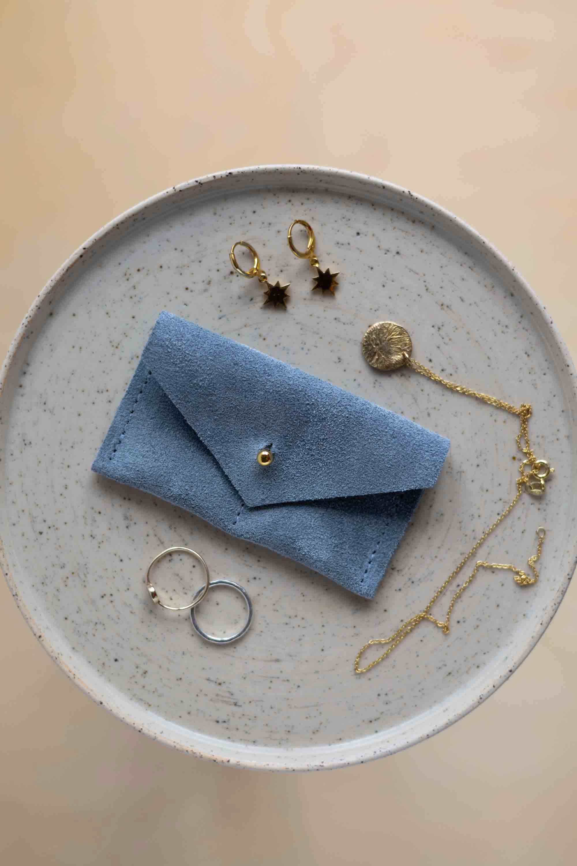 A top-down view of a dusky blue suede jewellery pouch with a gold coloured fastening, displayed on a round speckled ceramic plate alongside gold and silver jewellery pieces on a warm beige background.
