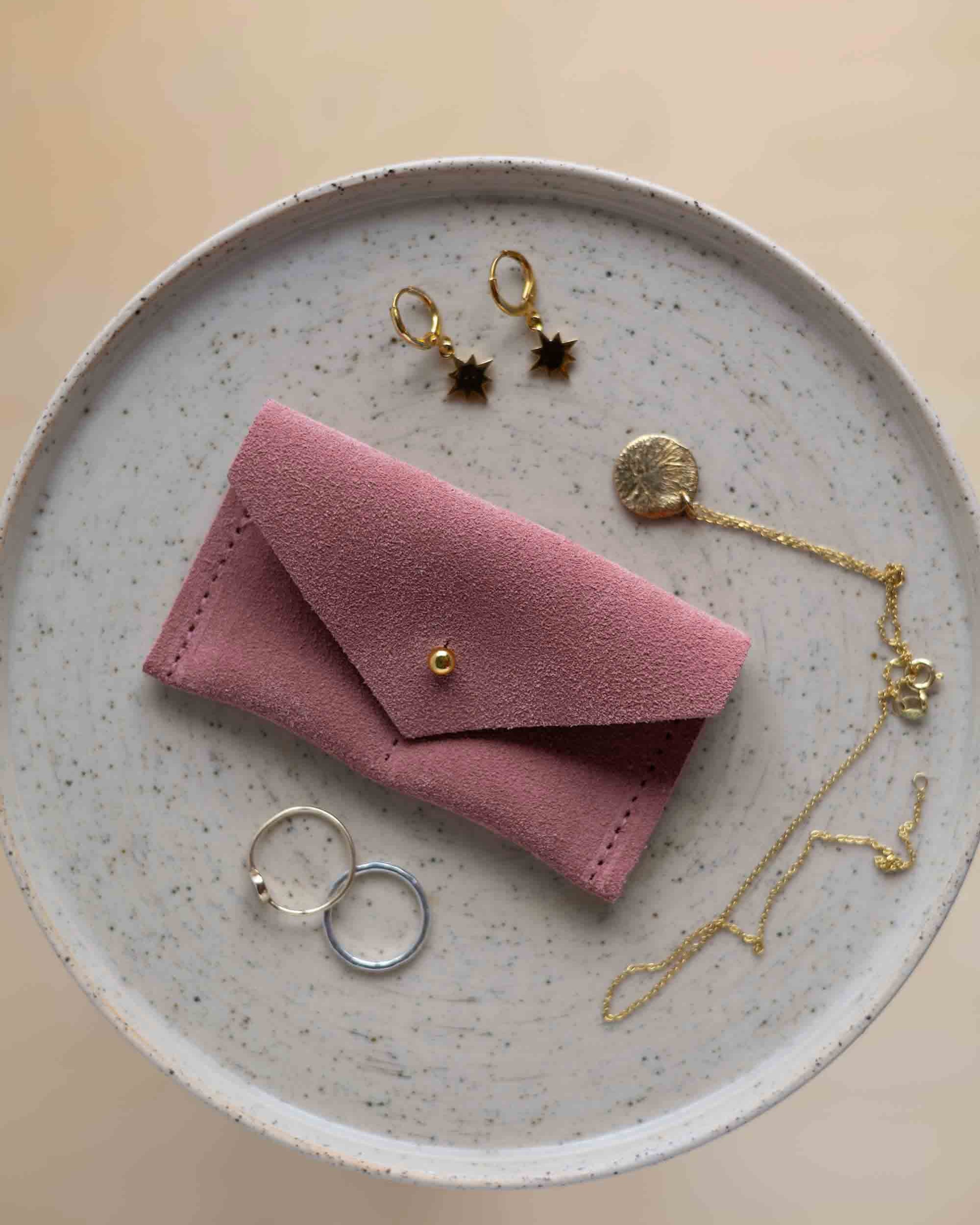 A top-down view of a dusky pink suede jewellery pouch with a gold coloured fastening, displayed on a round speckled plate alongside gold and silver jewellery pieces on a warm beige background.