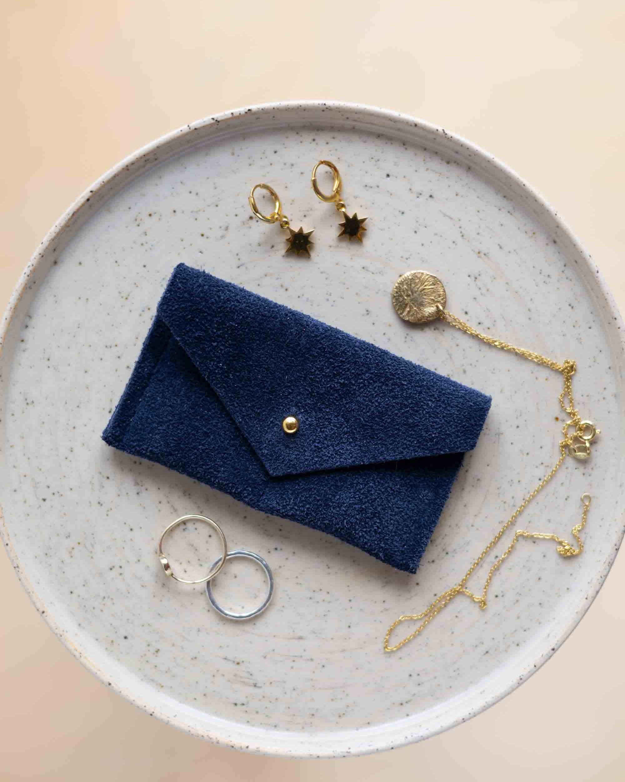 A top-down view of a midnight navy suede jewellery pouch with a gold coloured fastening, displayed on a round speckled ceramic plate alongside gold and silver jewellery pieces on a warm beige background.