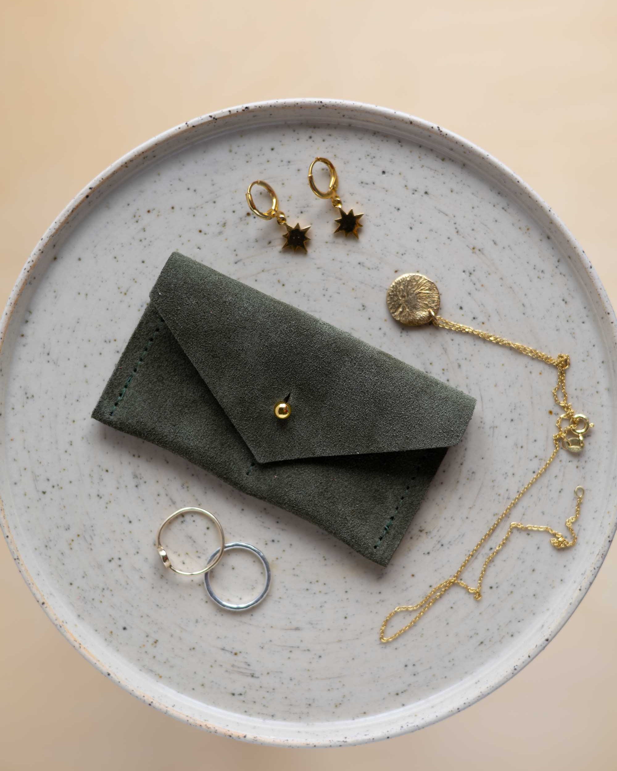 A top-down view of a soft olive green suede jewellery pouch with a gold coloured fastening, displayed on a round speckled ceramic plate alongside old and silver jewellery pieces on a warm beige background.