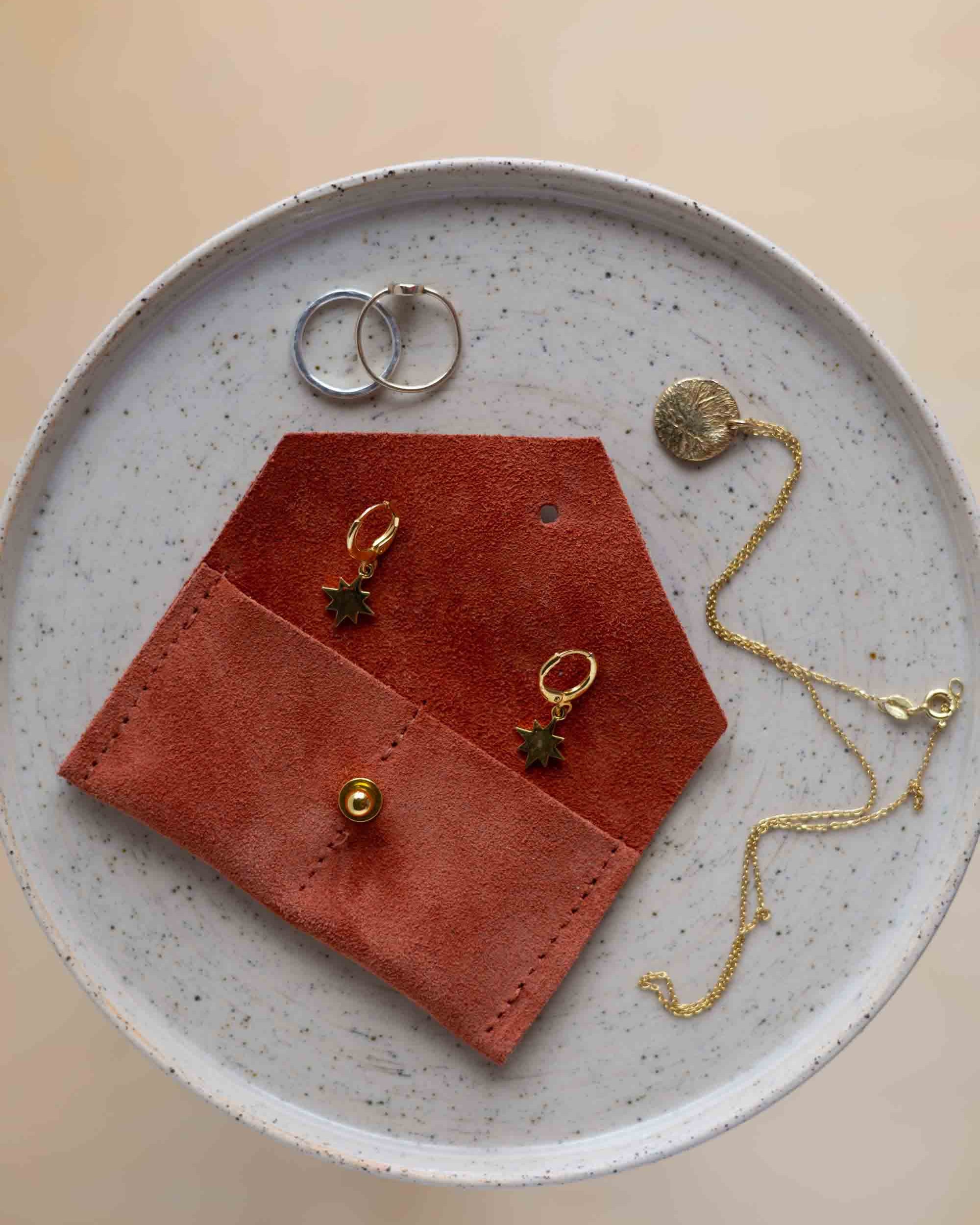 A top-down view of a terracotta coloured suede jewellery pouch with a gold coloured fastening, displayed on a round speckled ceramic plate alongside gold and silver jewellery pieces on a warm beige background.