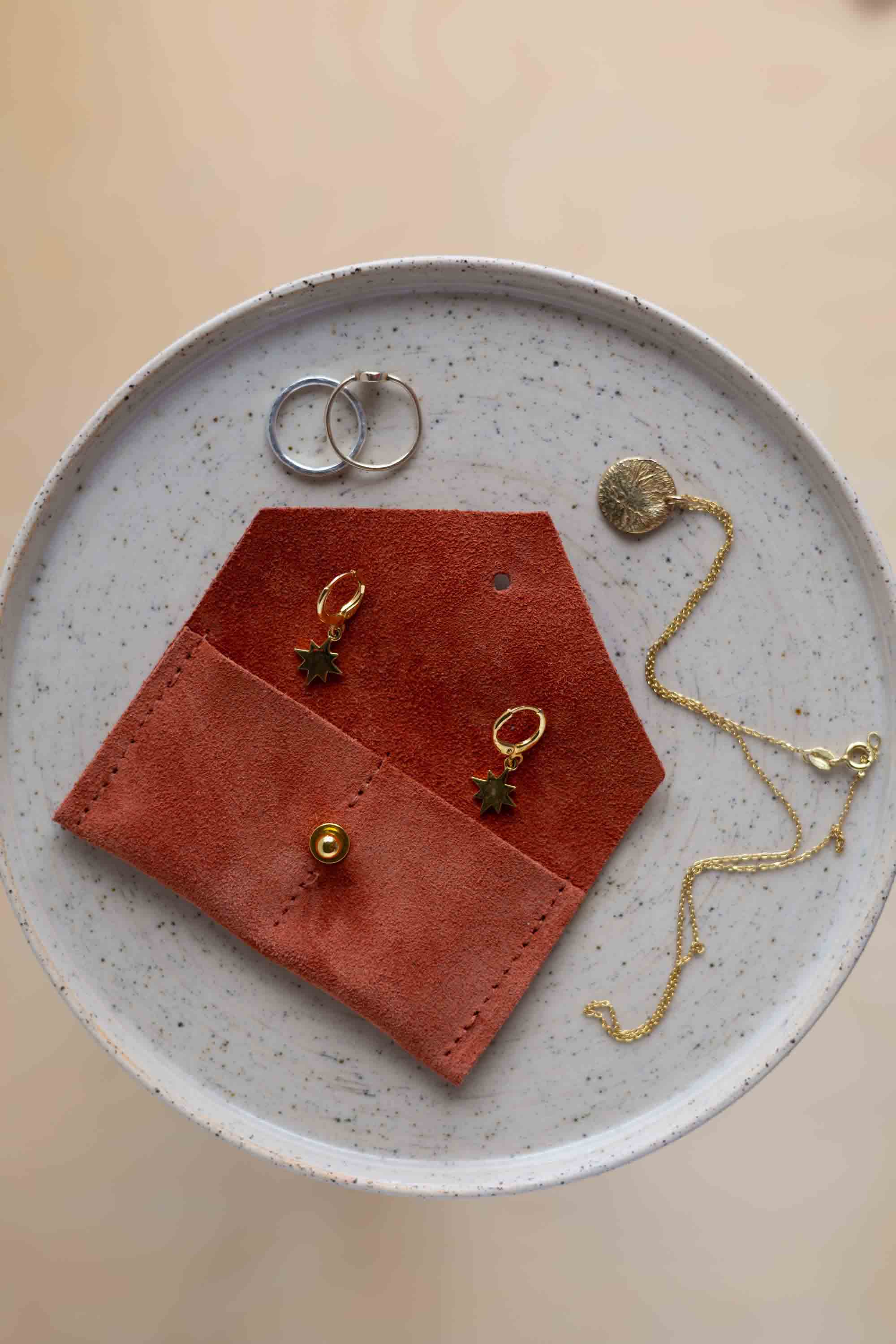A top-down view of a terracotta coloured suede jewellery pouch with a gold coloured fastening, displayed on a round speckled ceramic plate alongside gold and silver jewellery pieces on a warm beige background.