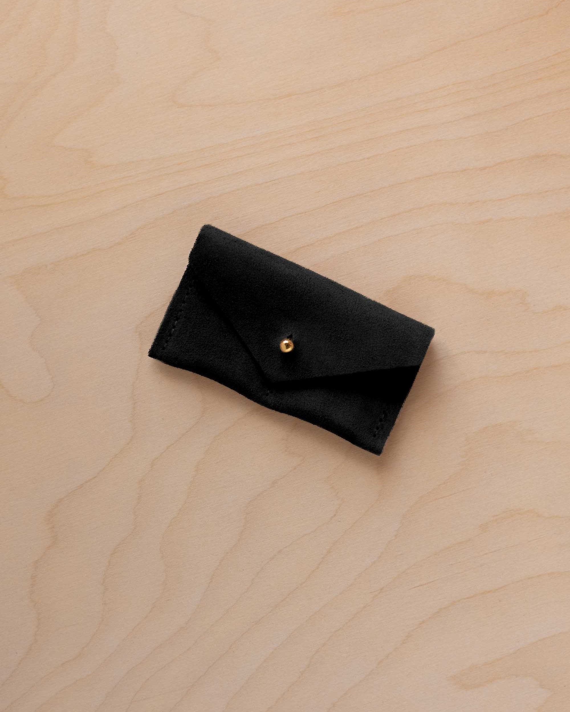 A small black coloured suede jewellery pouch with a gold button fastening, resting on a light wood grain surface.