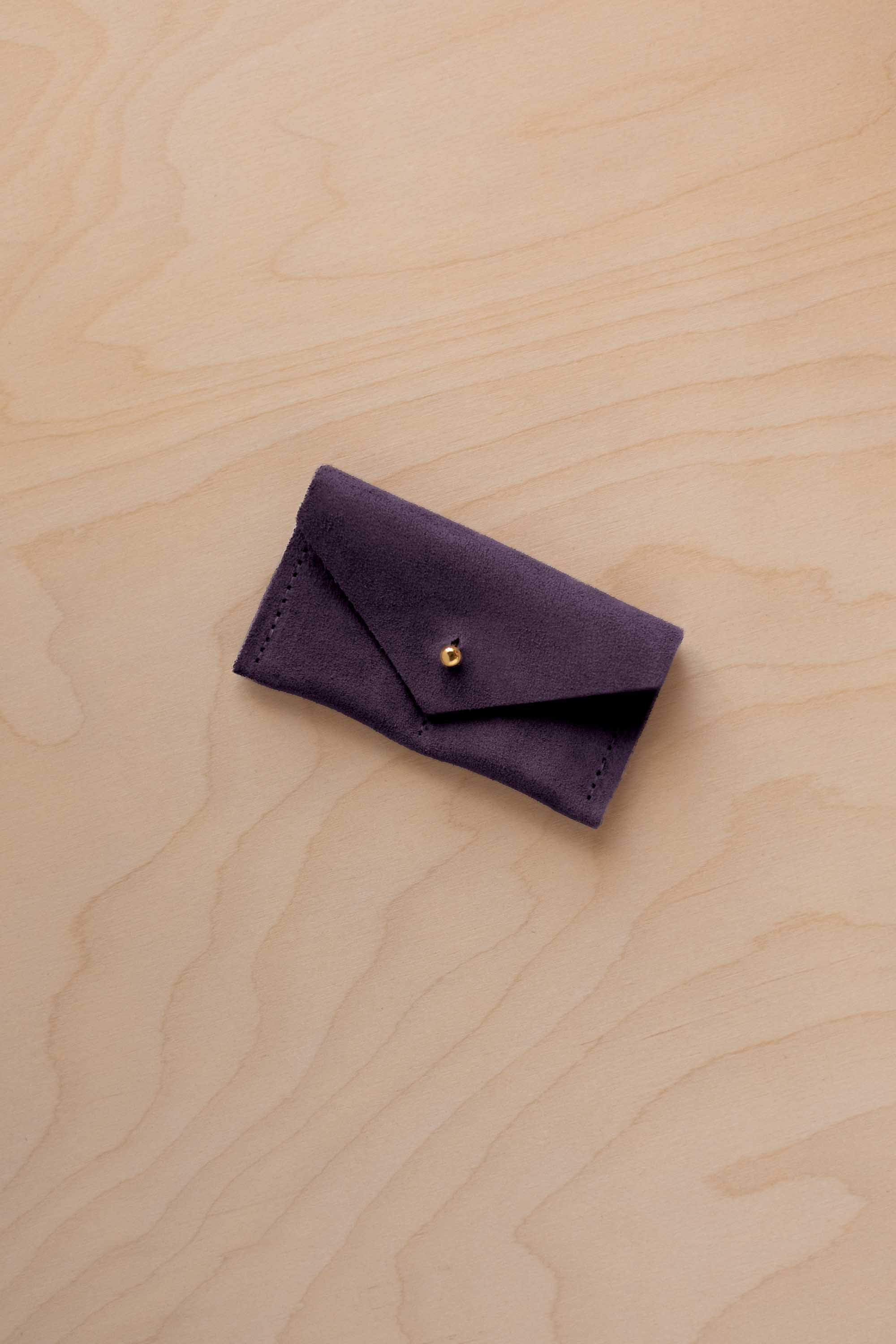 A small blackcurrant coloured suede jewellery pouch with a gold coloured fastening, resting on a light wood grain surface.