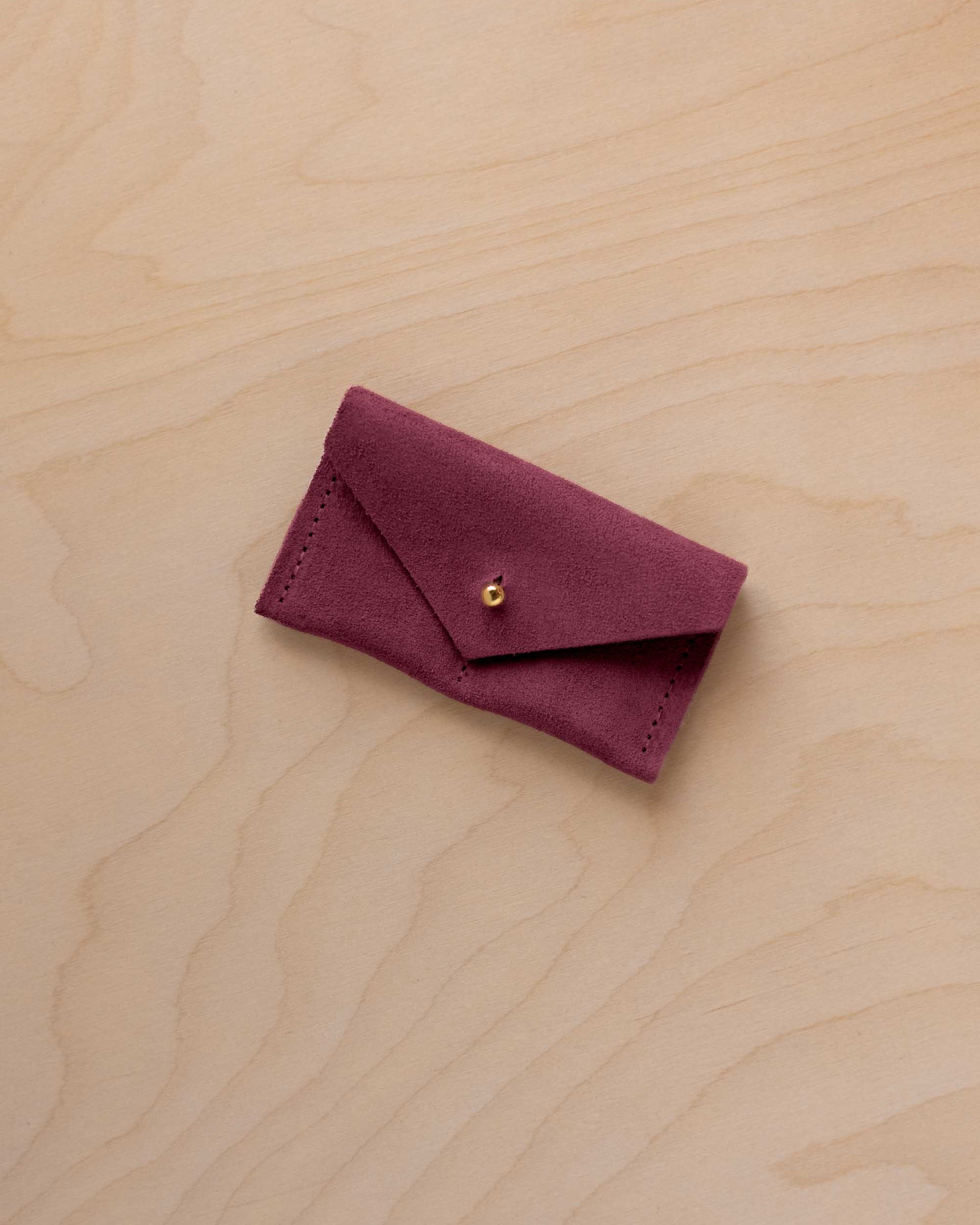 A small burgundy coloured suede jewellery pouch with a gold coloured fastening, resting on a light wood grain surface.