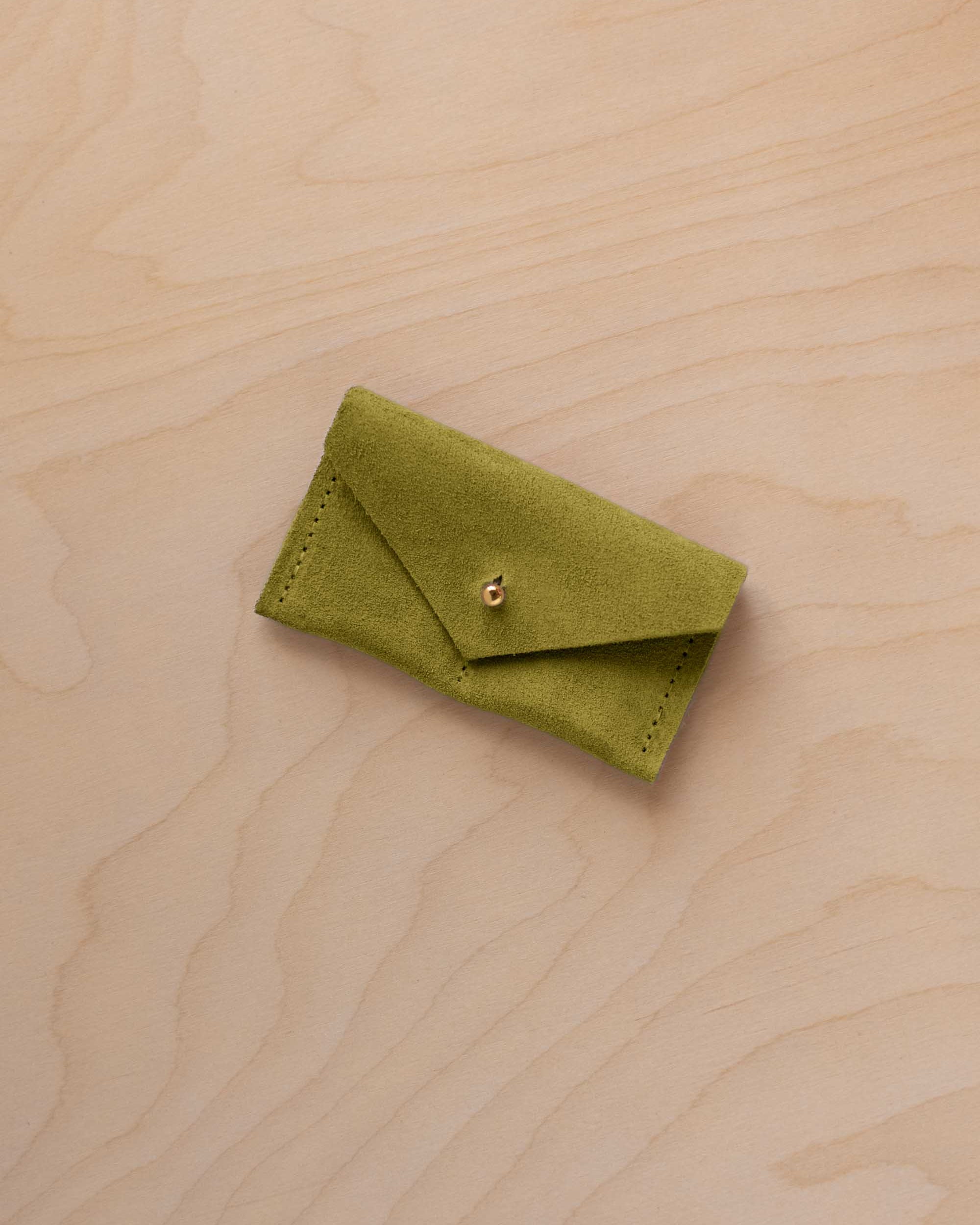 A small chartreuse green coloured suede jewellery pouch with a gold button, resting on a light wood grain surface.