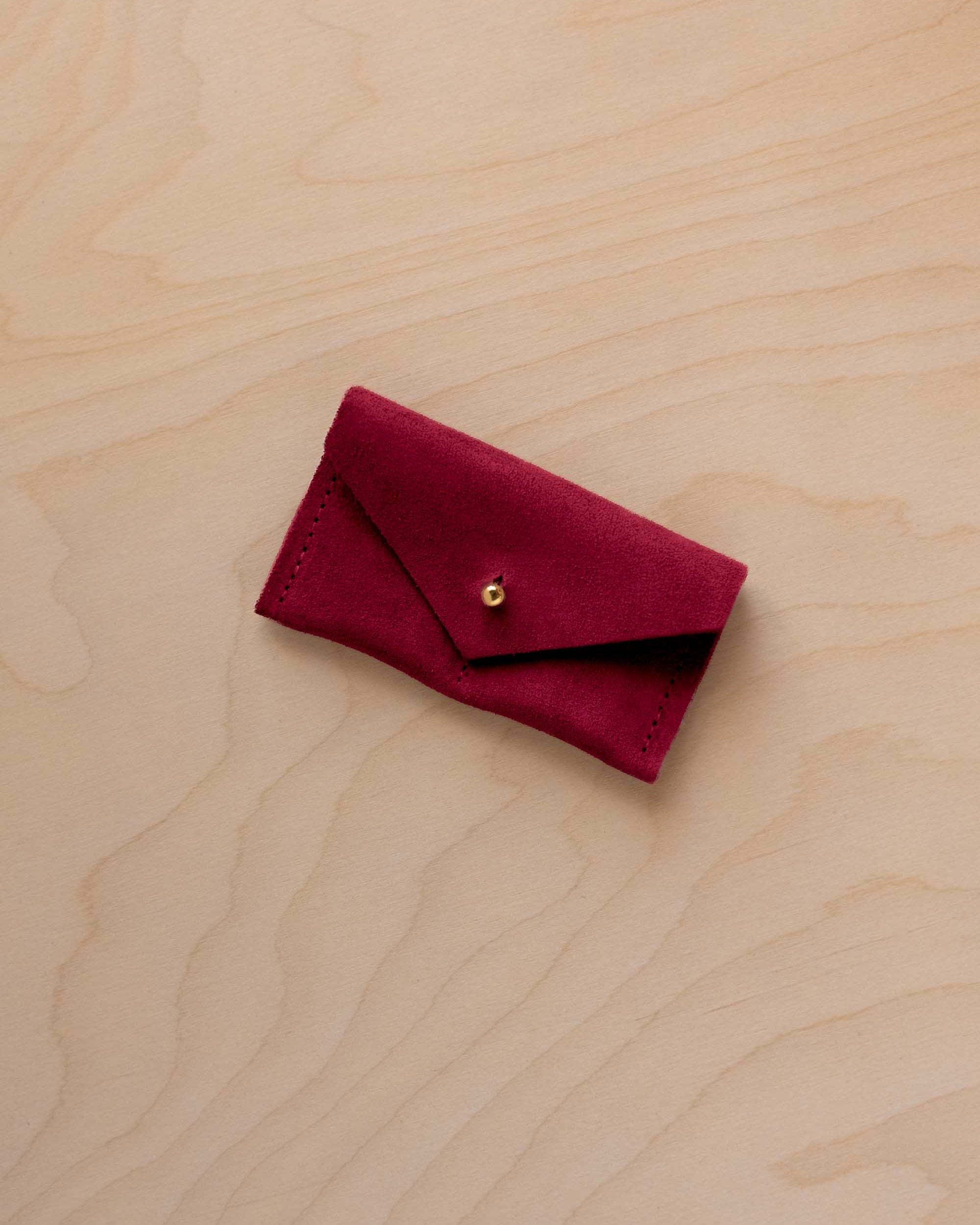 A small crimson red coloured suede jewellery pouch with a gold coloured fastening, resting on a light wood grain surface.