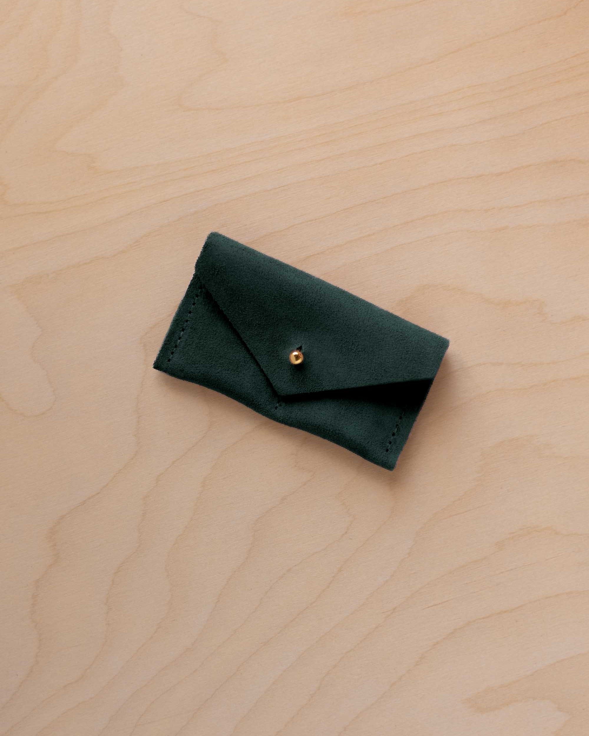A small deep forest coloured suede jewellery pouch with a gold button, resting on a light wood grain surface.
